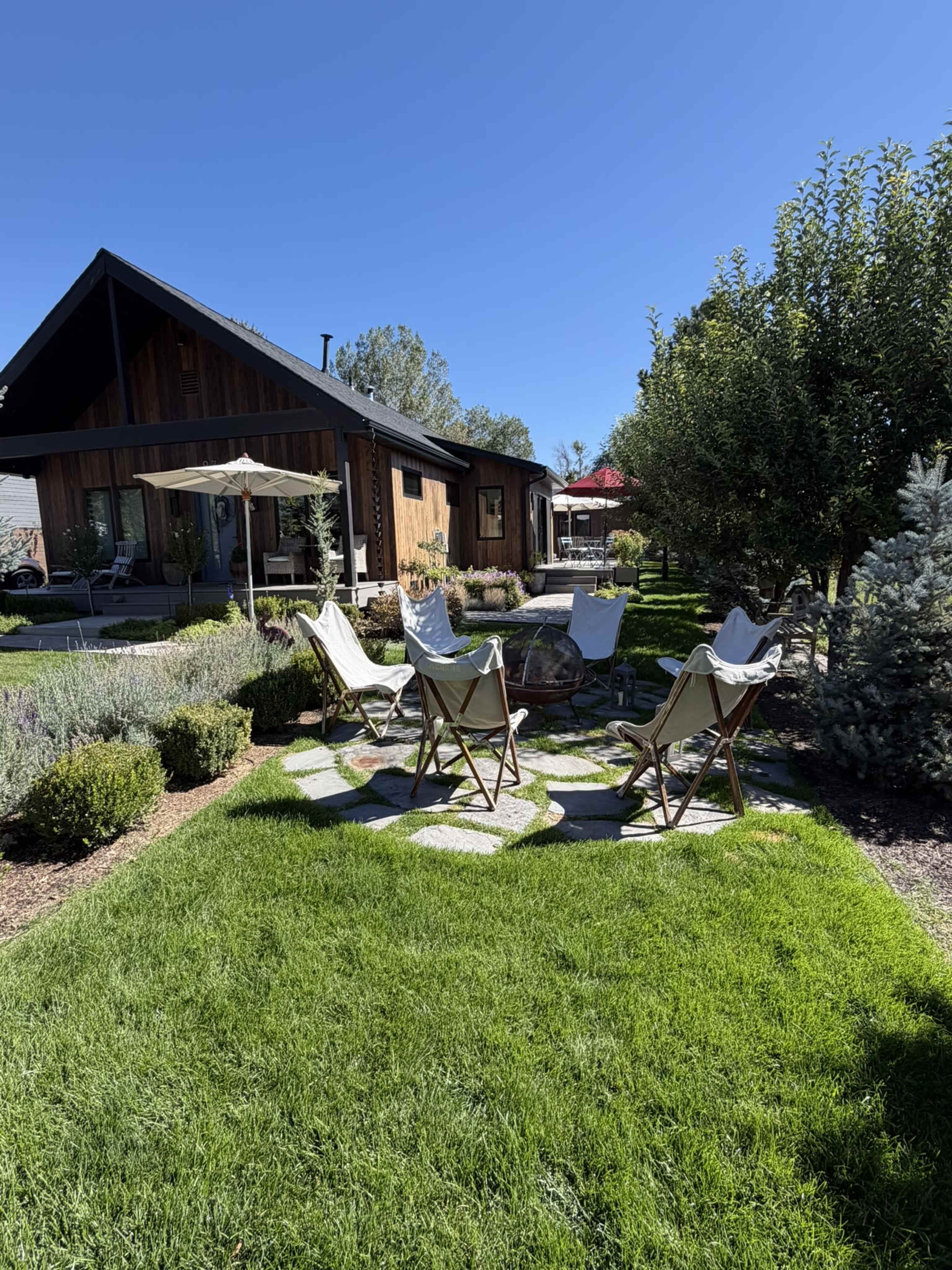 The image shows a backyard with a stone patio surrounded by four folding chairs, grass, and various shrubs, with a house in the background.