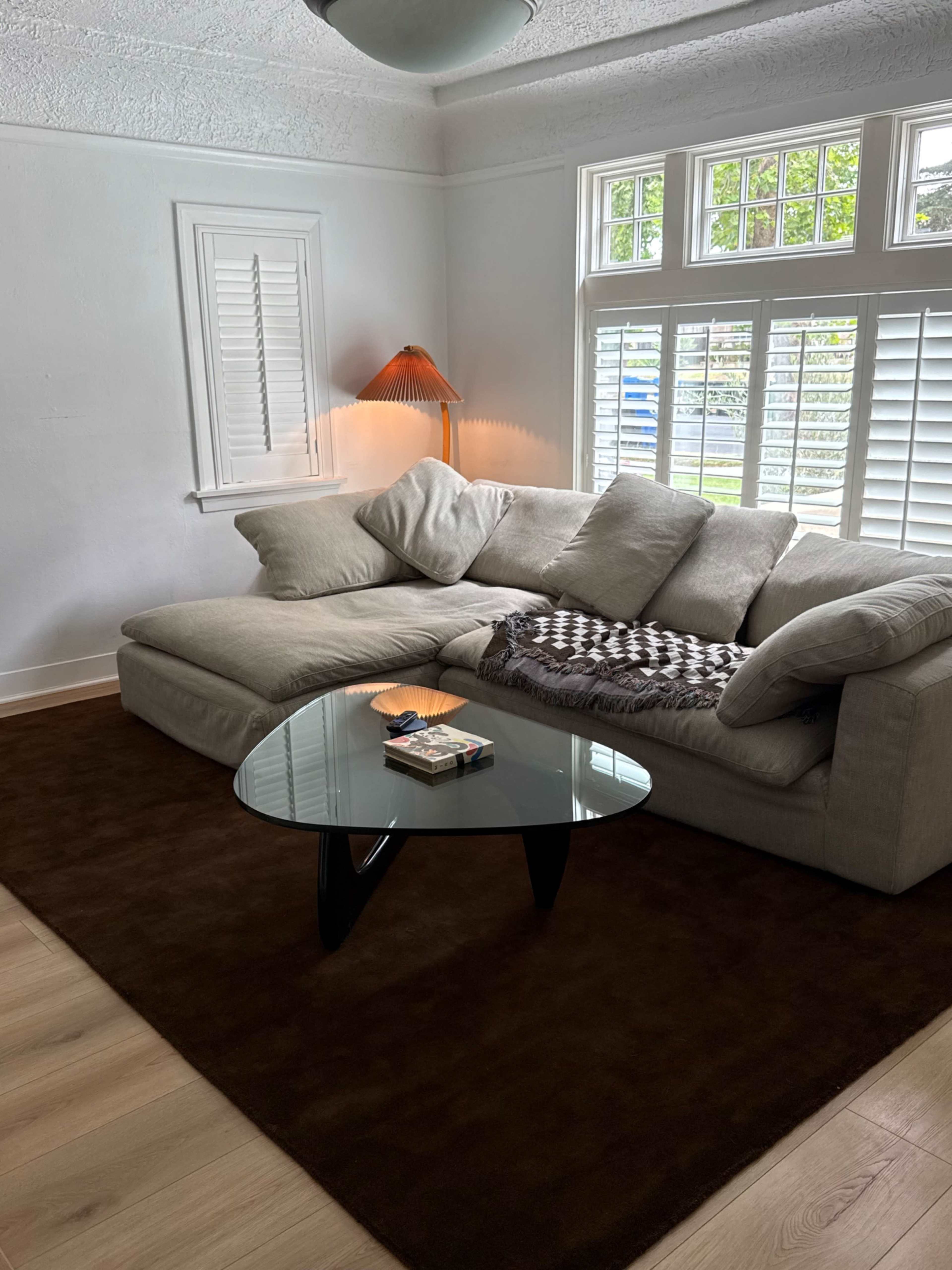 The image shows a light-colored sectional sofa with scattered cushions, a glass coffee table, and a lamp in a well-lit room featuring window shutters and wooden flooring.