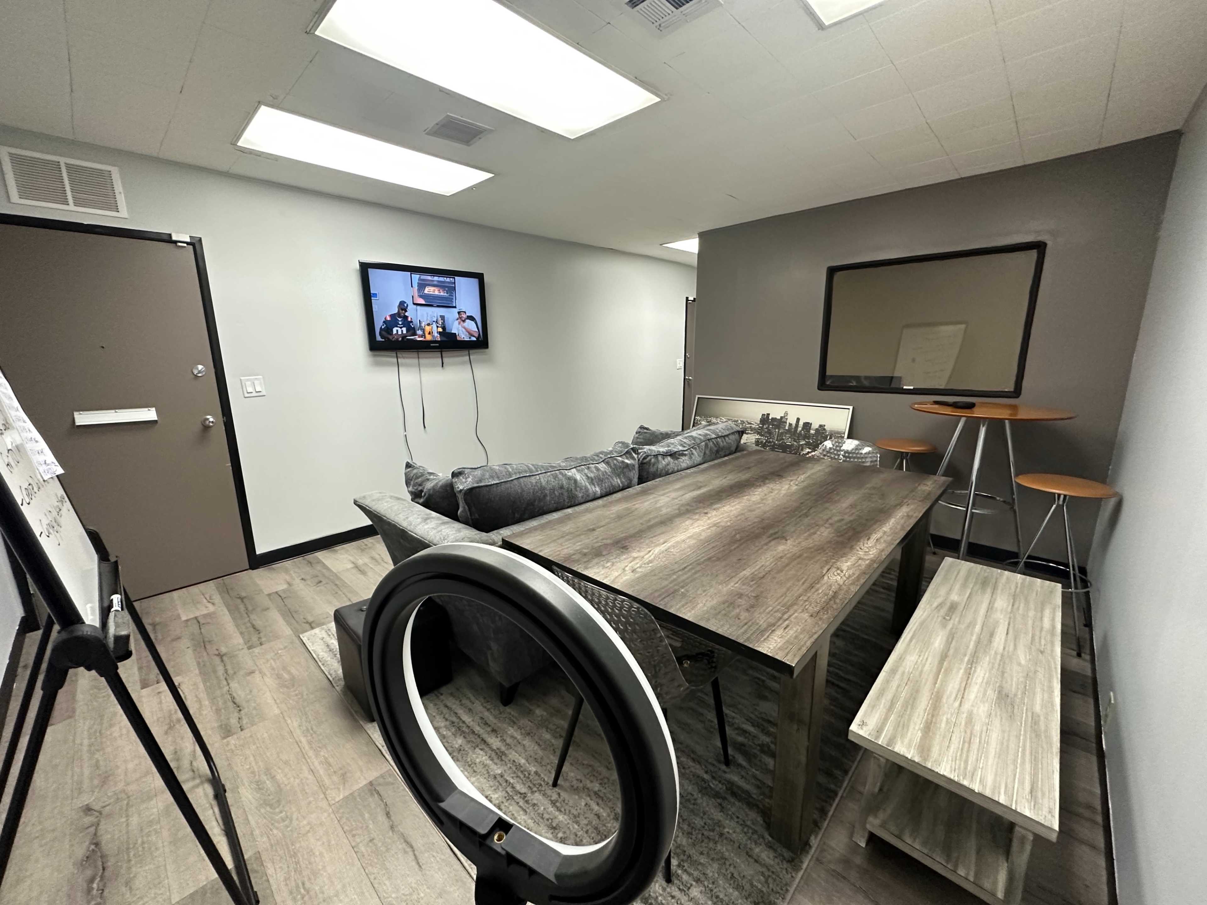 A modern conference room equipped with a wooden table, a large TV on the wall, a mirror, and seating arrangements including a sofa and a stool.