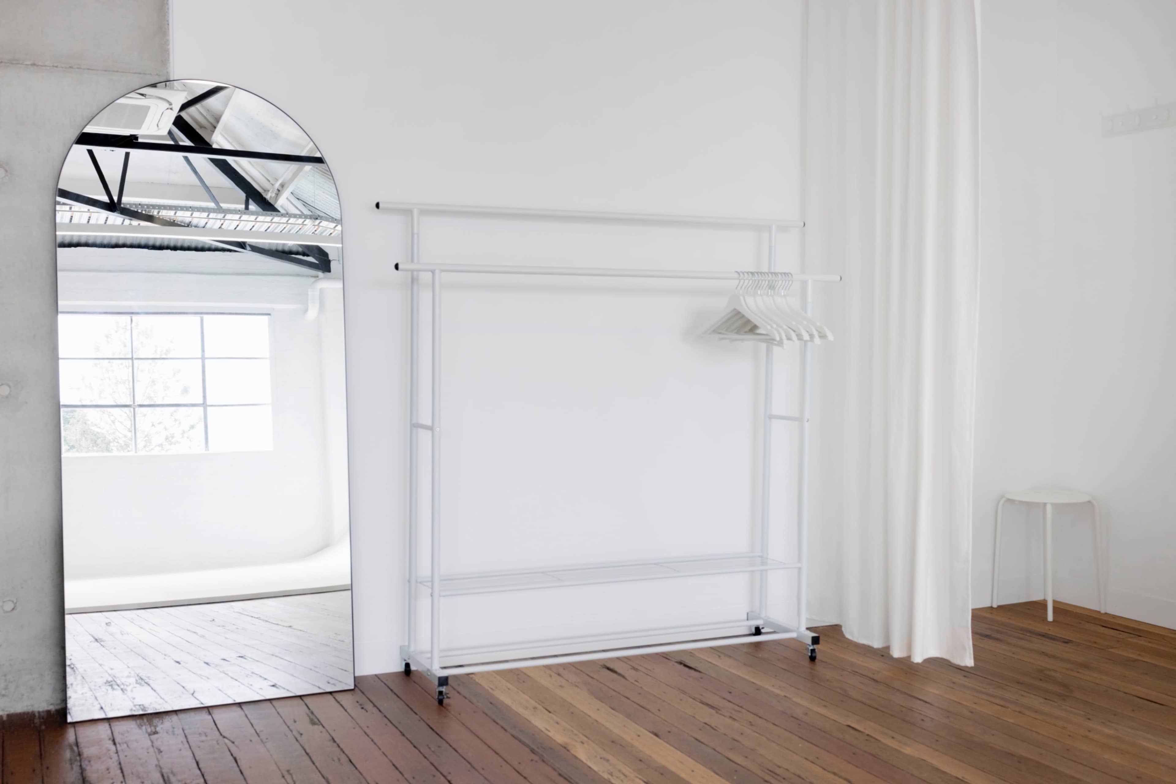 A minimalist room featuring a large arch-shaped mirror, a freestanding clothing rack with hangers, and white curtains beside a wooden floor.