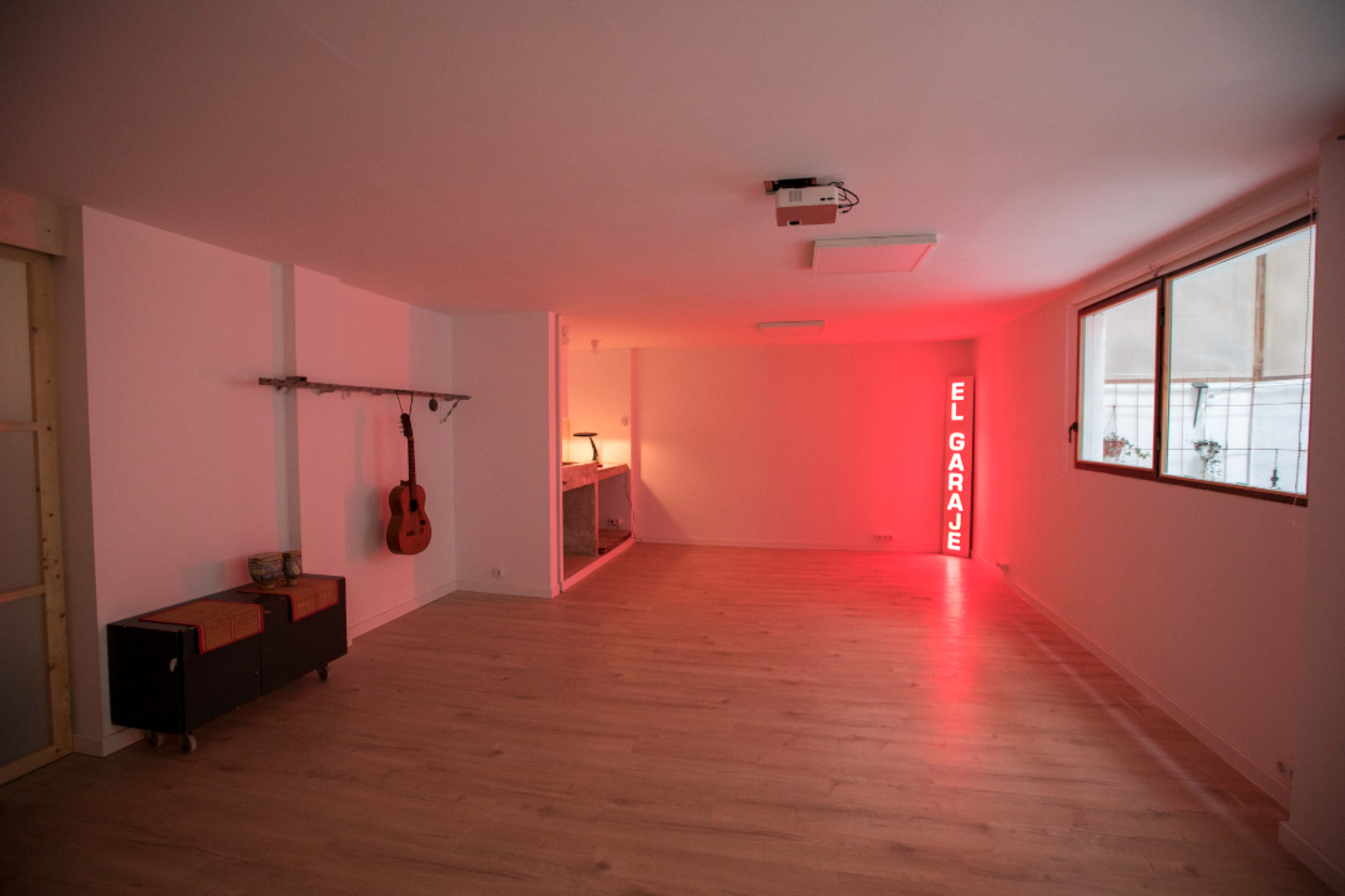 A spacious interior room with light wooden flooring, a guitar hung on a wall, a countertop area, and a neon sign reading "EL GARAGE."