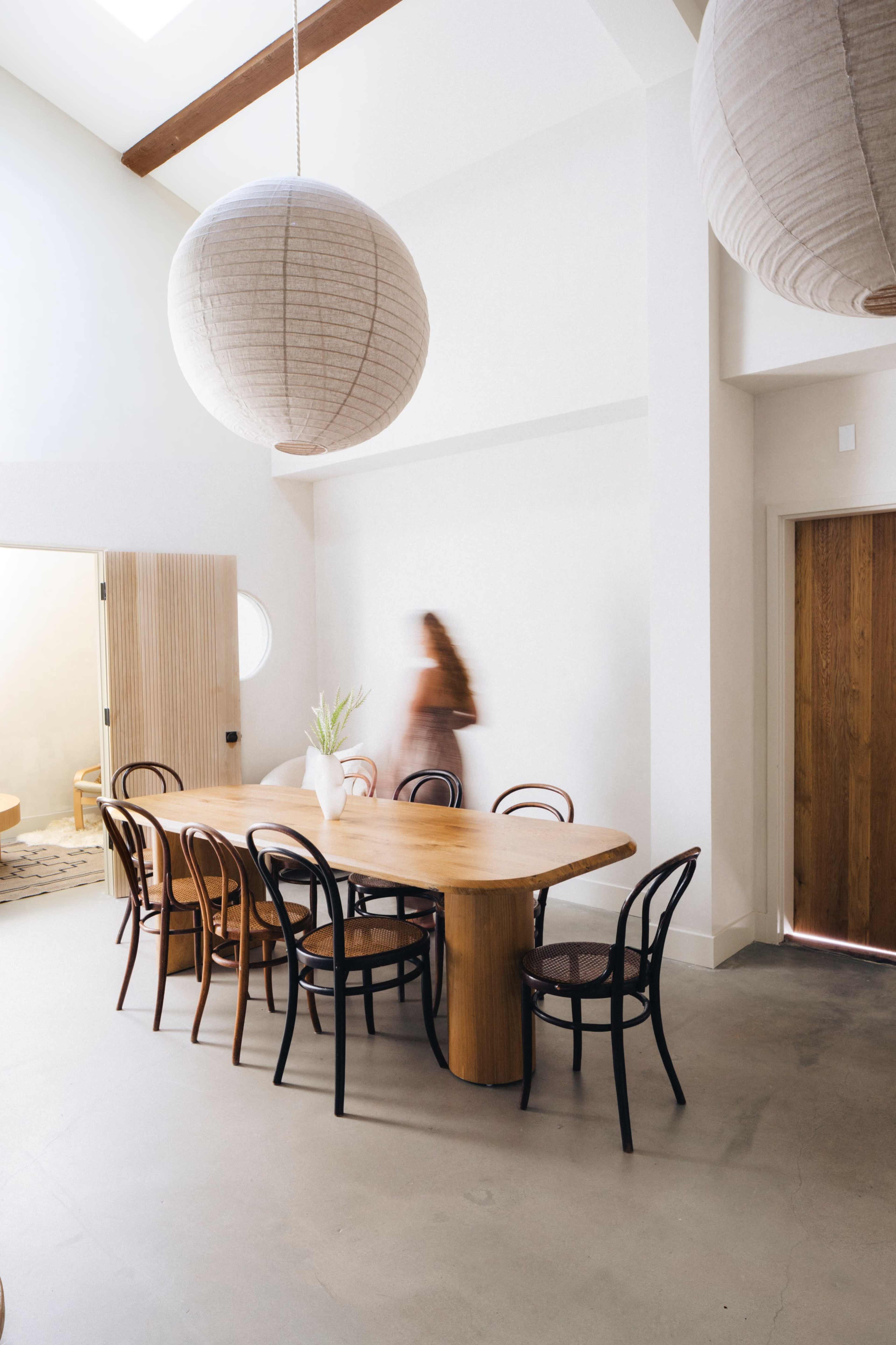 A spacious dining area features a large wooden table surrounded by several black and natural chairs, with pendant lights hanging above.