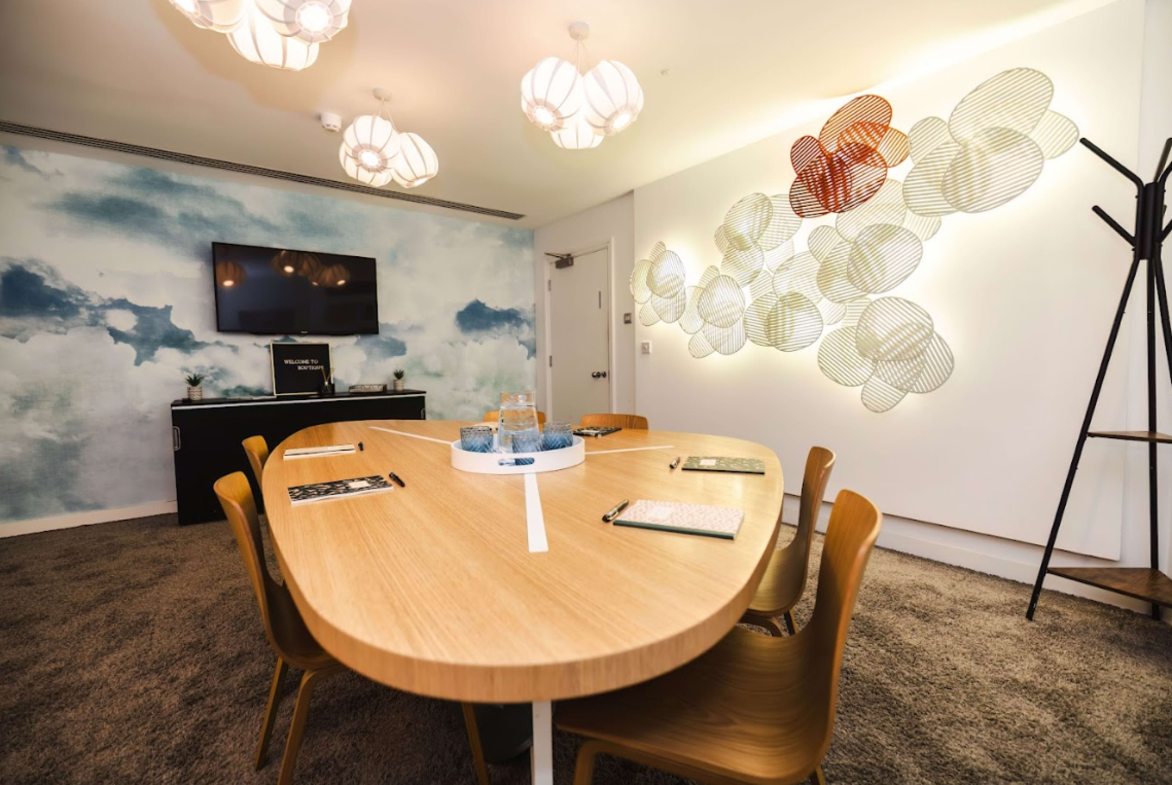 A modern conference room with a long wooden table, chairs, a TV mounted on the wall, and a decorative mural featuring clouds.