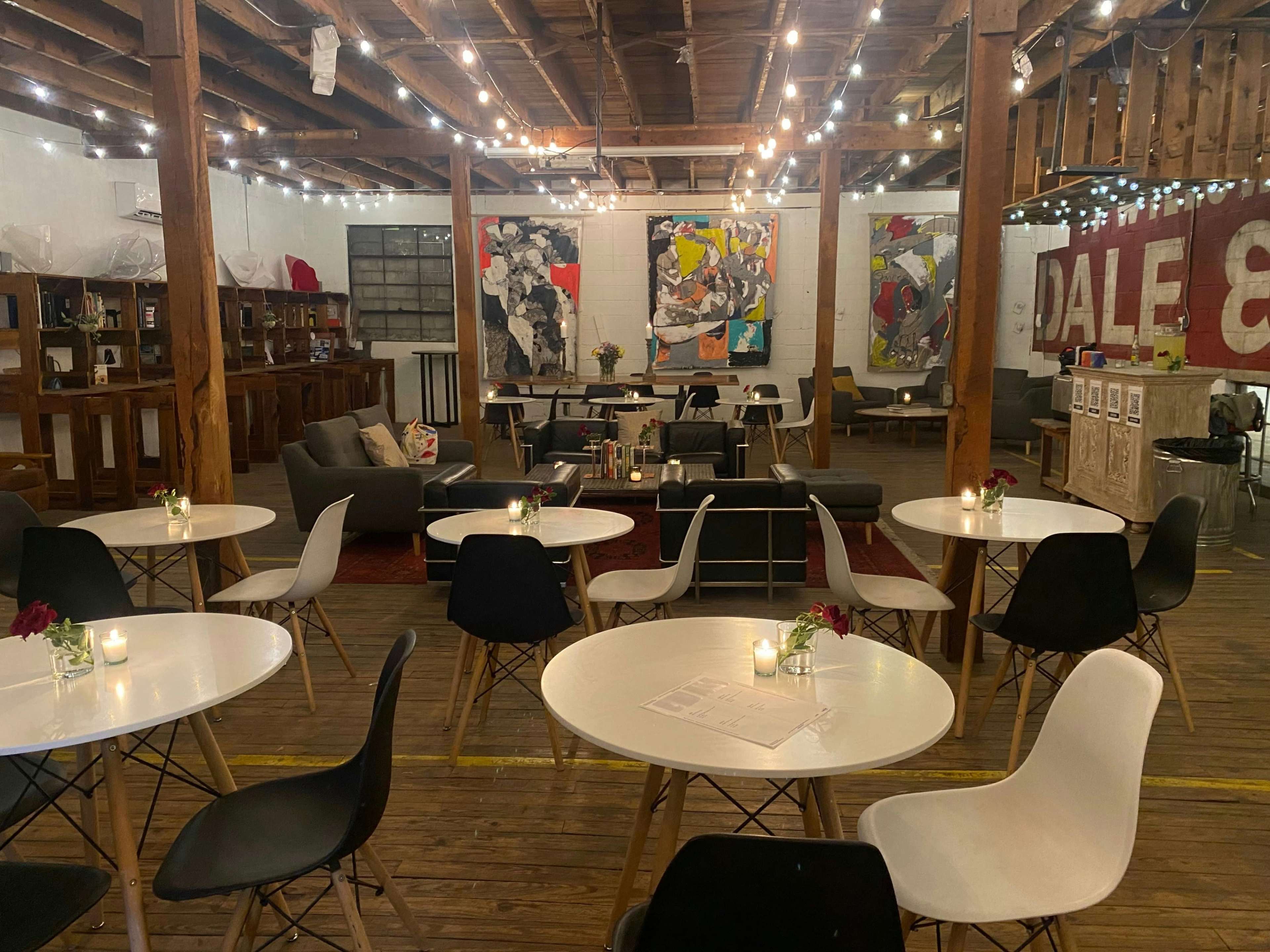 A spacious cafe or event space with round tables, chairs, and artwork on the walls, illuminated by string lights hanging from the ceiling.