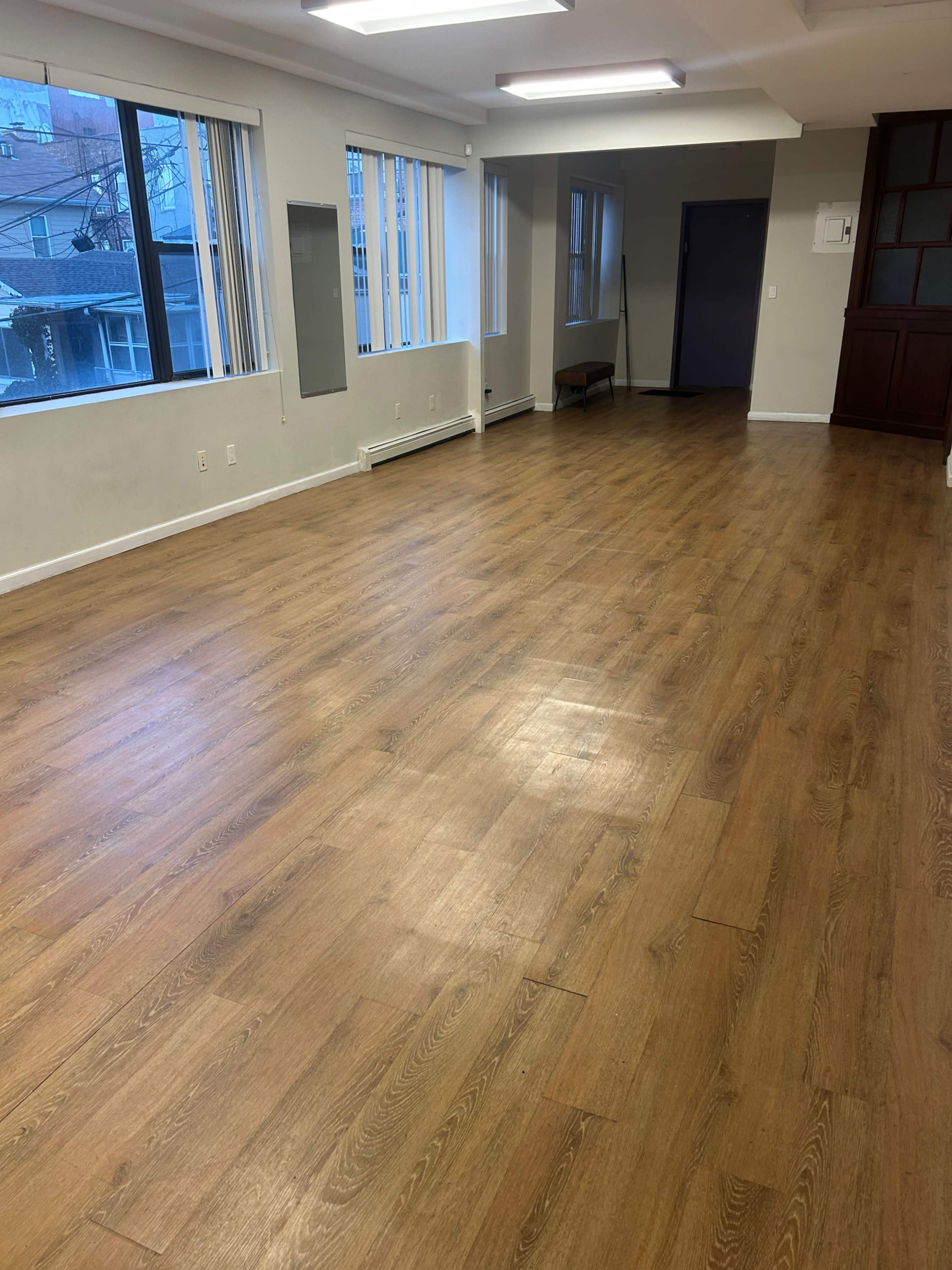 The image shows a spacious room with wooden flooring, large windows on one side, and minimal furnishings.