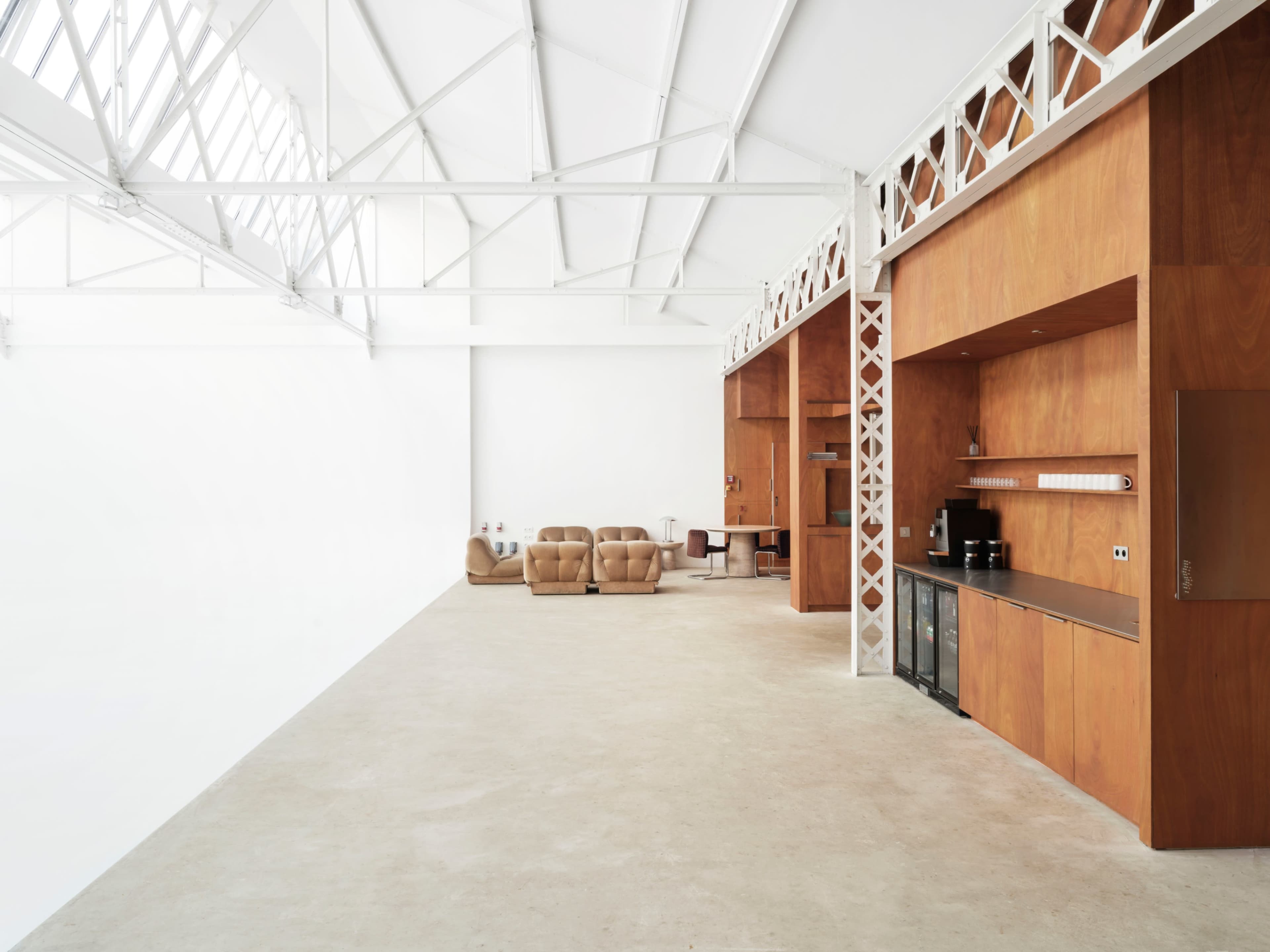 The image shows a spacious, modern interior featuring wooden walls, a kitchenette area, two brown armchairs, and an empty concrete floor.
