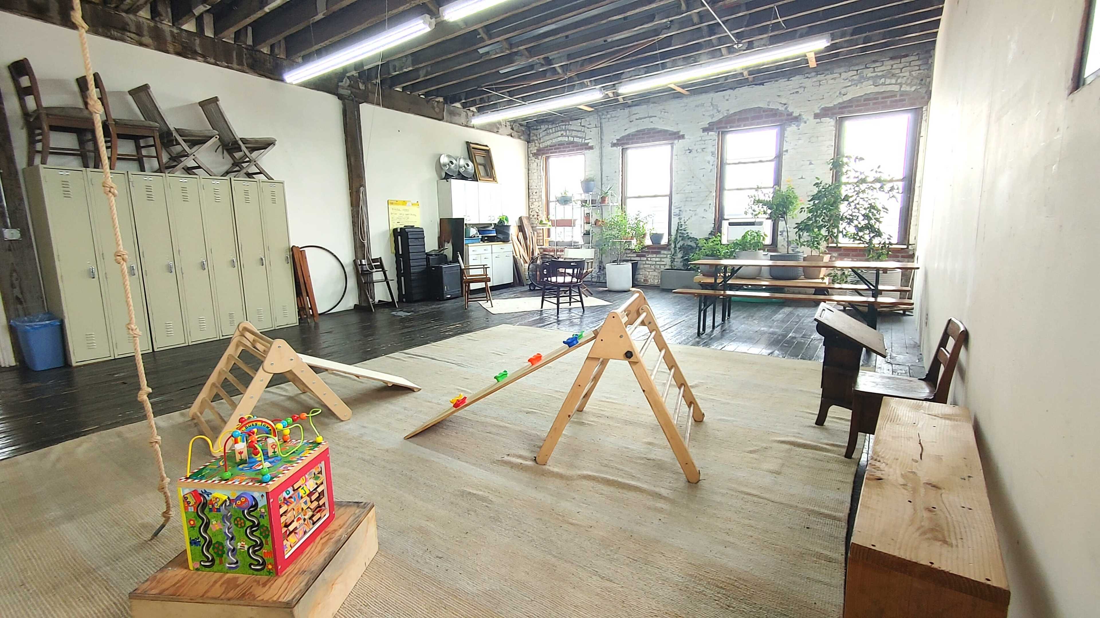 The image shows a spacious indoor play area featuring wooden climbing frames, a colorful activity cube, and storage lockers along one wall.