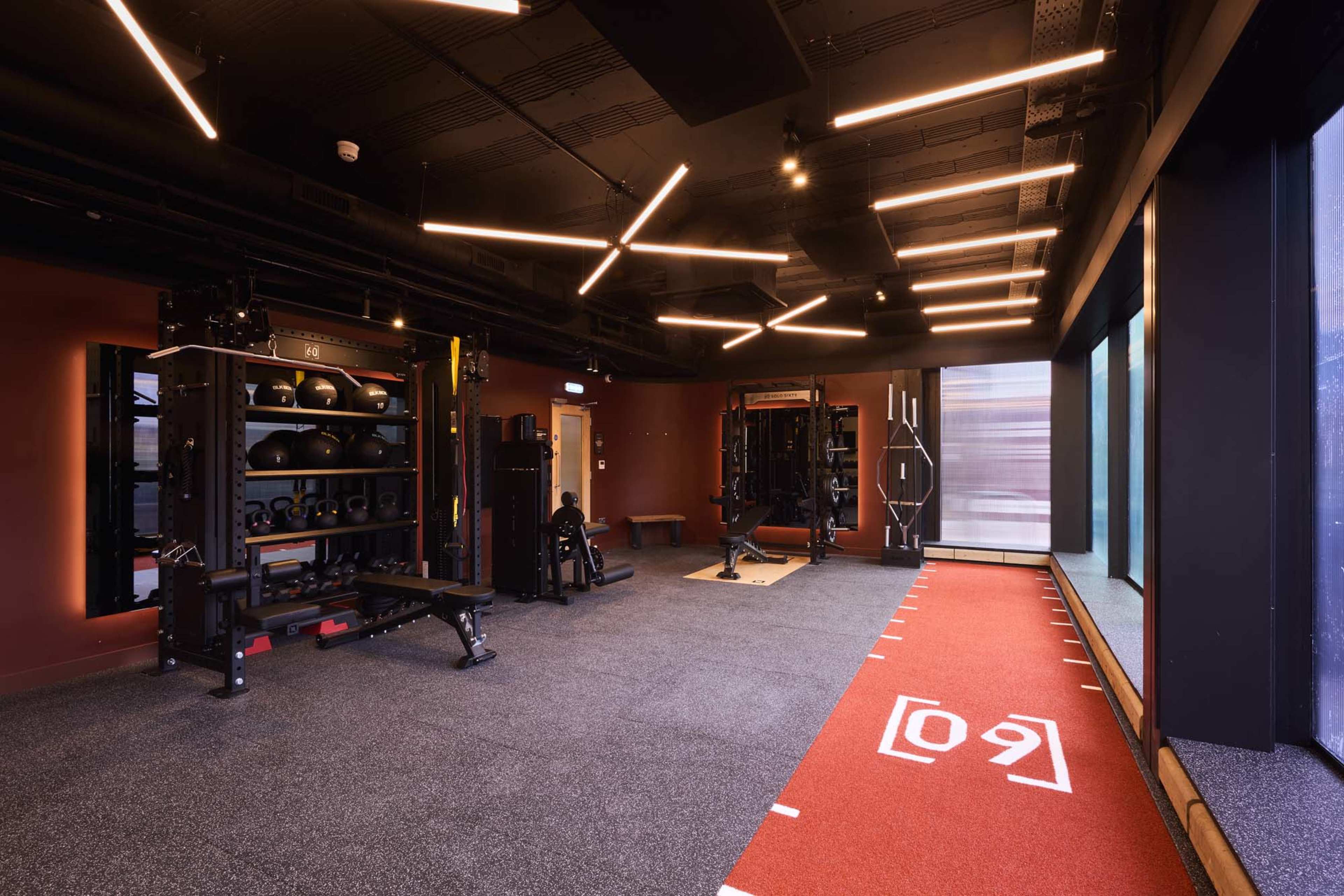 The image shows a modern gym interior featuring various fitness equipment, including weight racks and a training area with a red track.