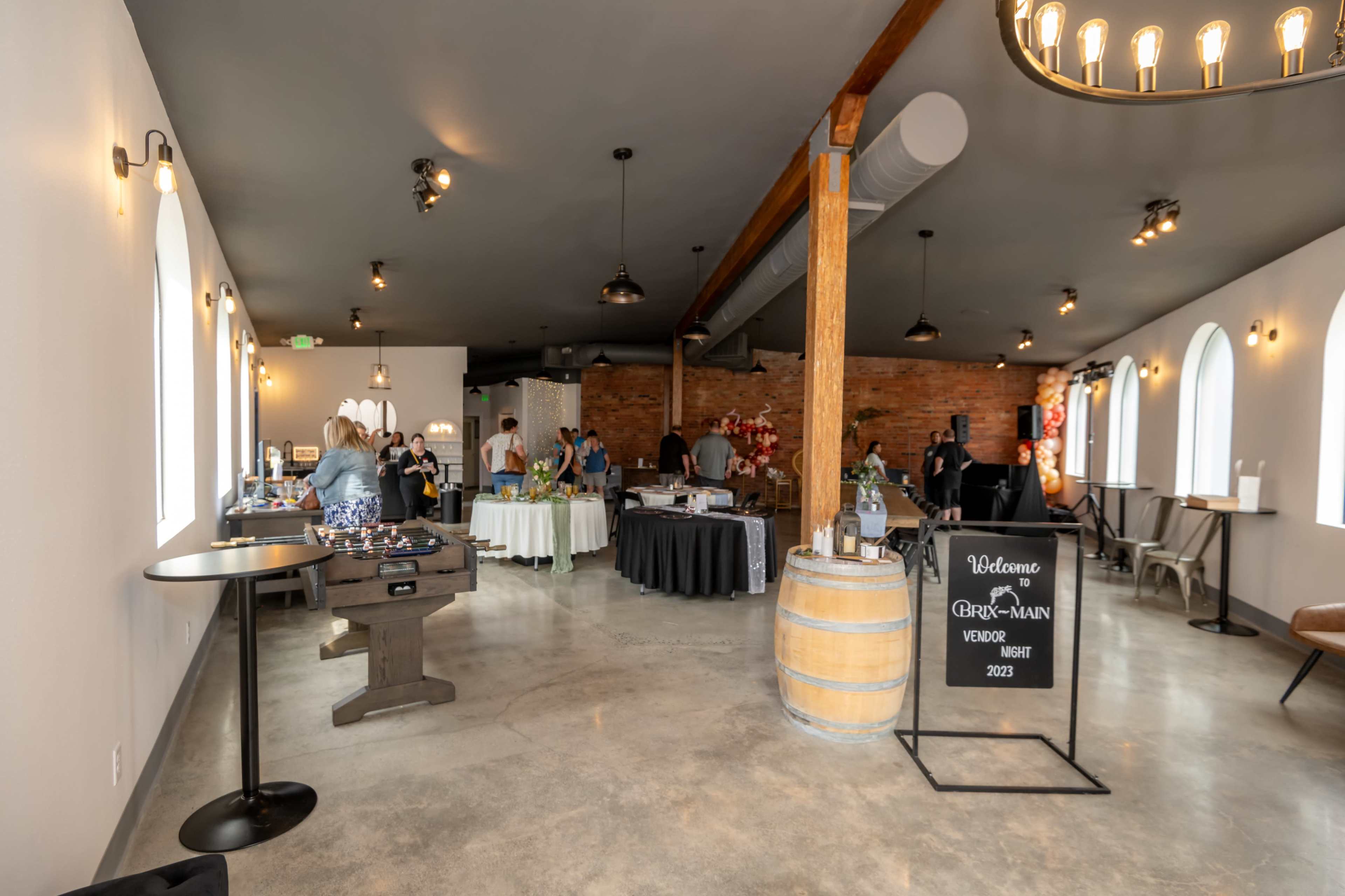 A spacious event venue features a mix of vendors, tables, and decor, with exposed brick walls and large windows.