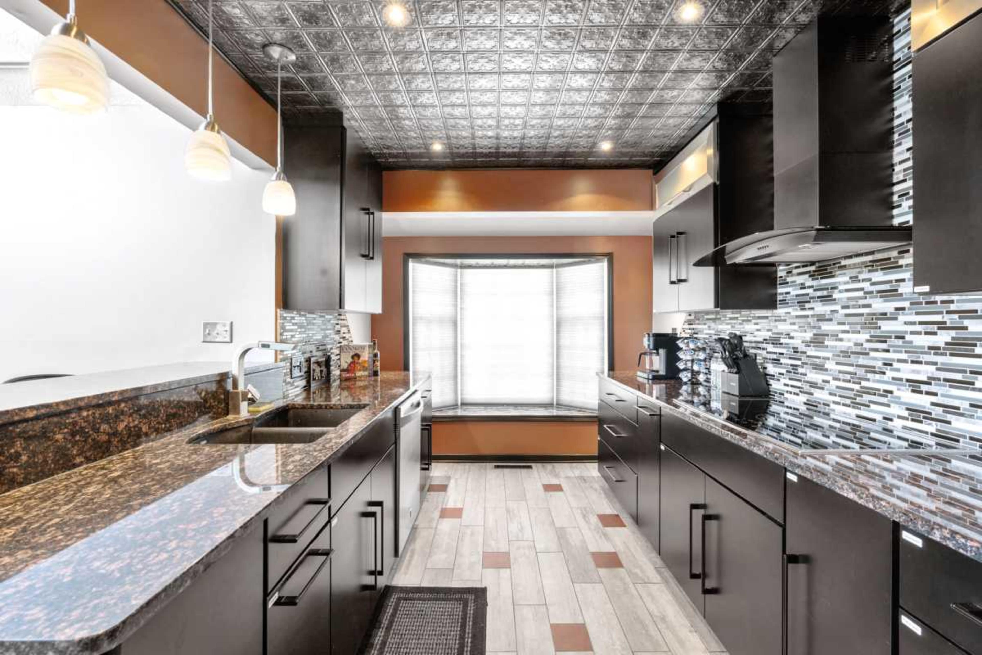The image shows a modern kitchen featuring dark cabinets, granite countertops, and a tiled backsplash, with abundant natural light coming from a large window.
