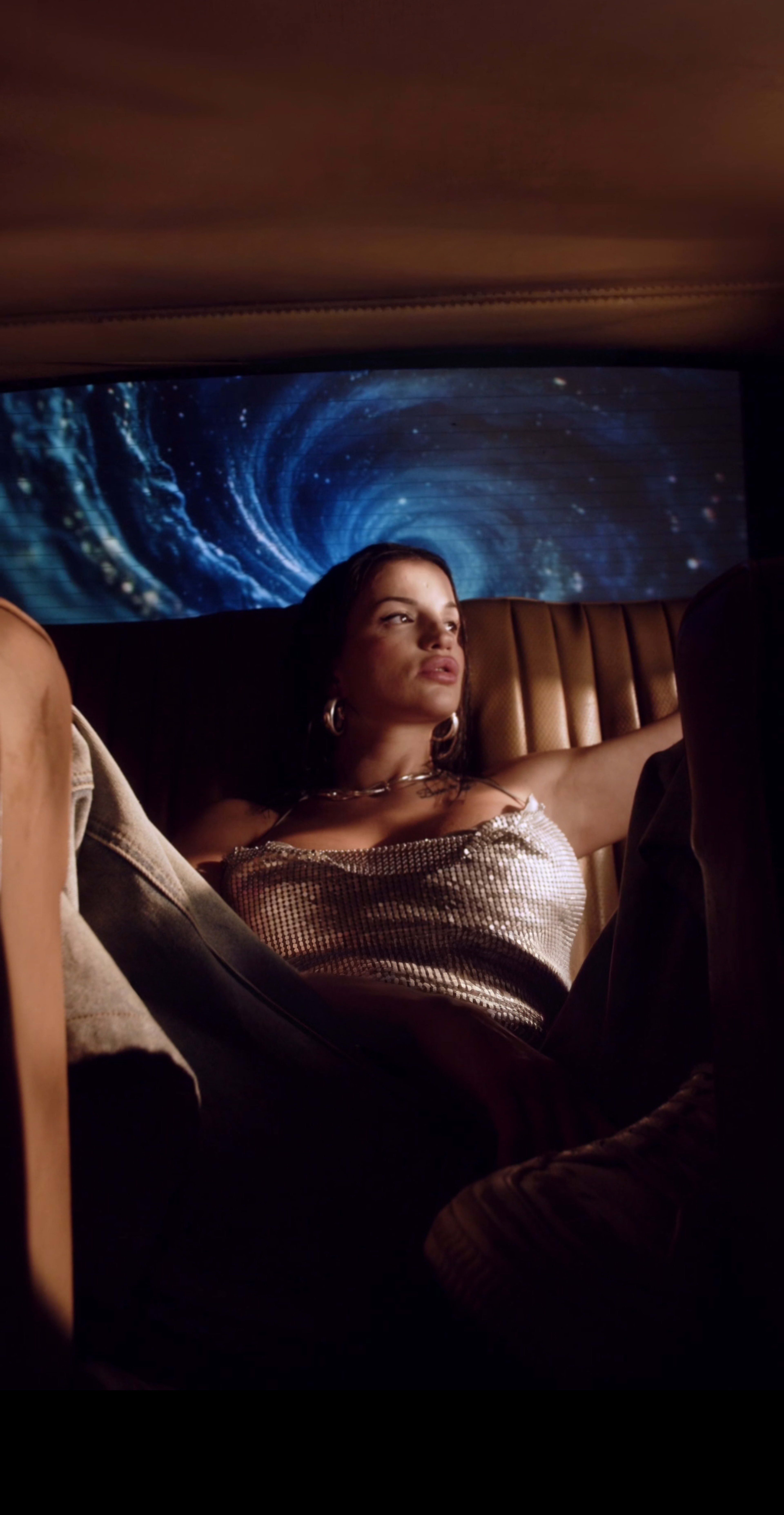 A woman lounges in the back seat of a car, with a swirling cosmic backdrop projected behind her.