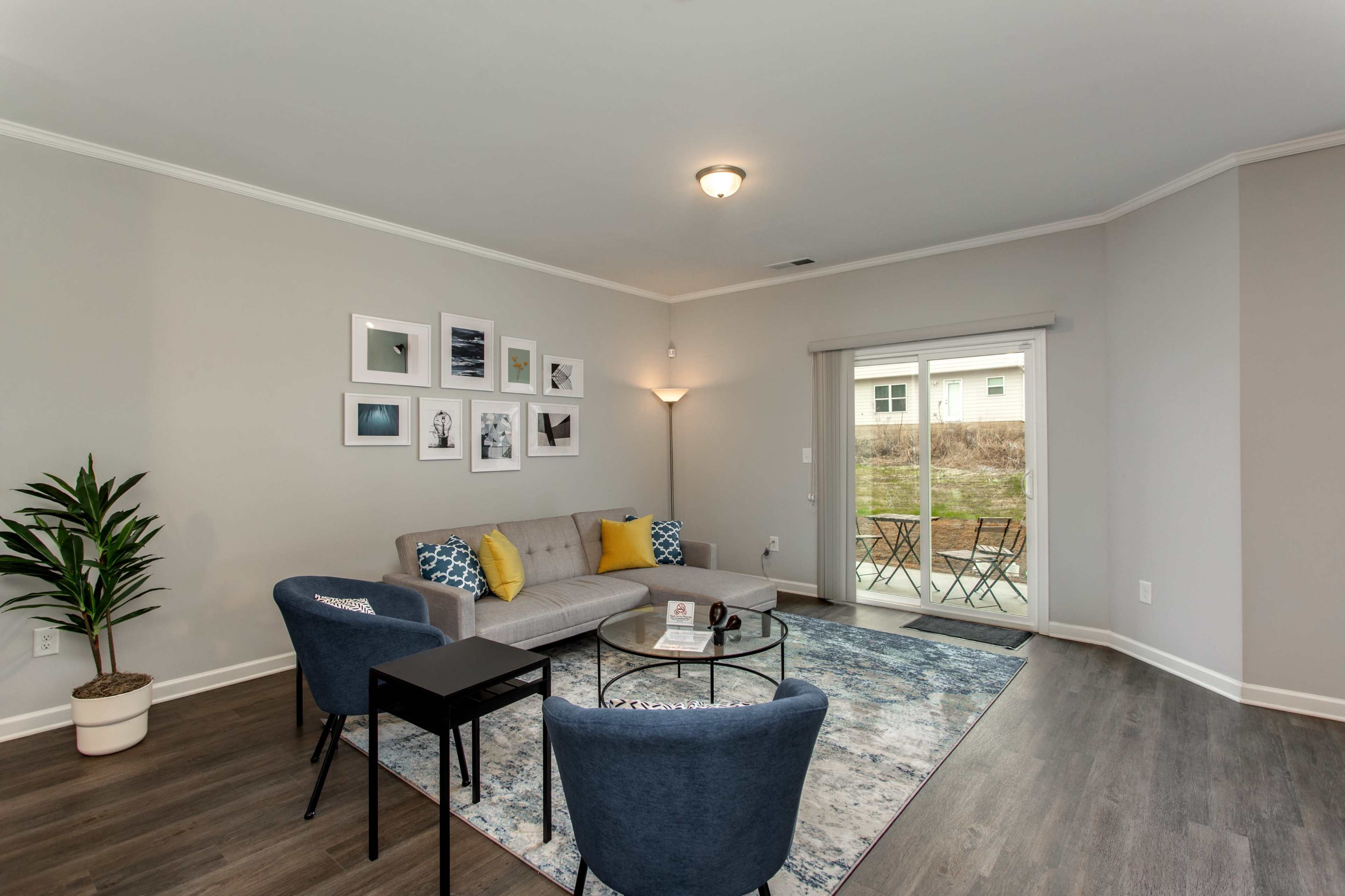 A modern living room with a gray sofa, two blue chairs, a small black table, and a sliding glass door leading to an outdoor seating area.