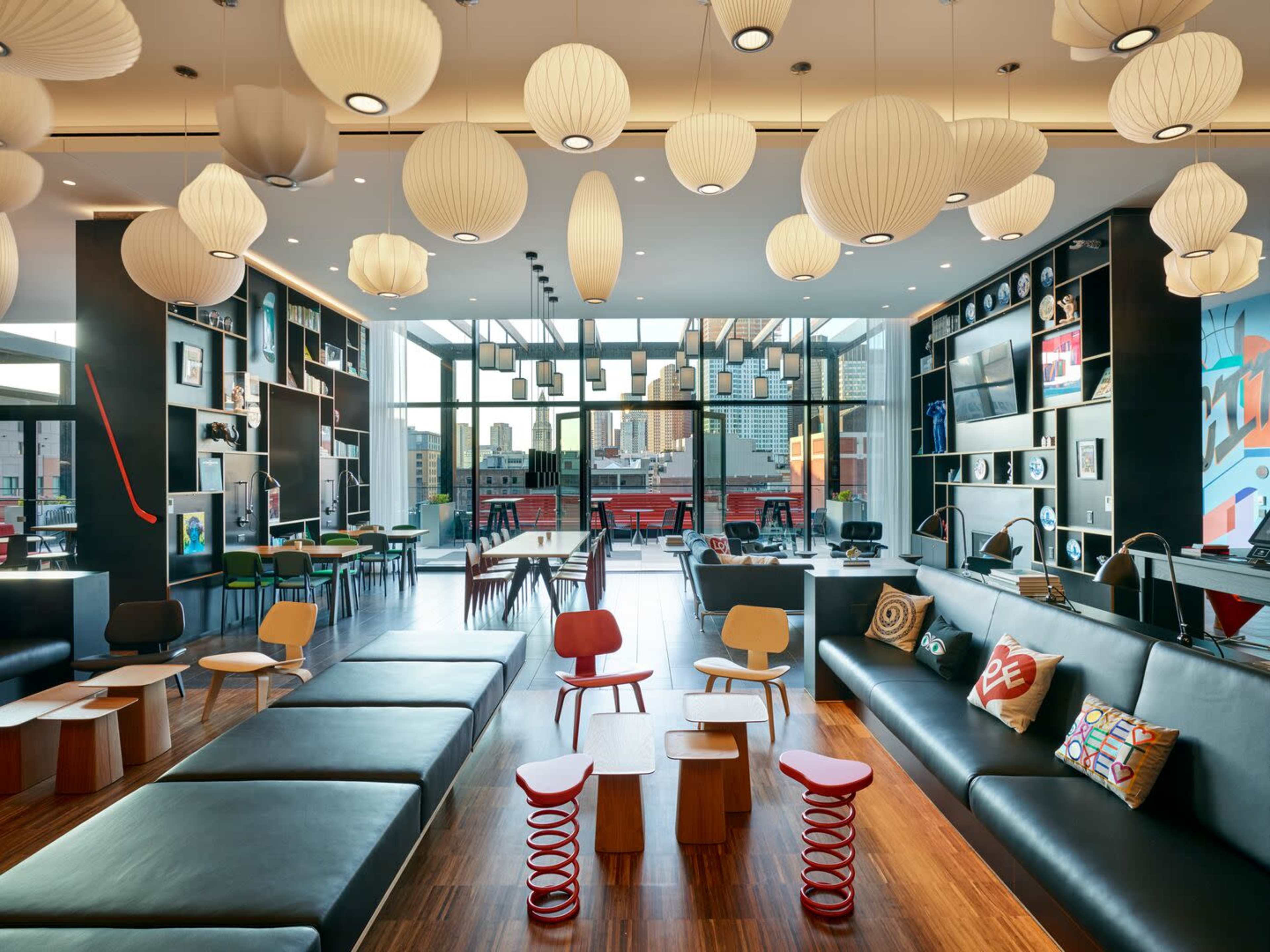 A modern lounge area with a mix of black sofas, colorful small chairs, and pendant lighting, featuring large windows that overlook a cityscape.