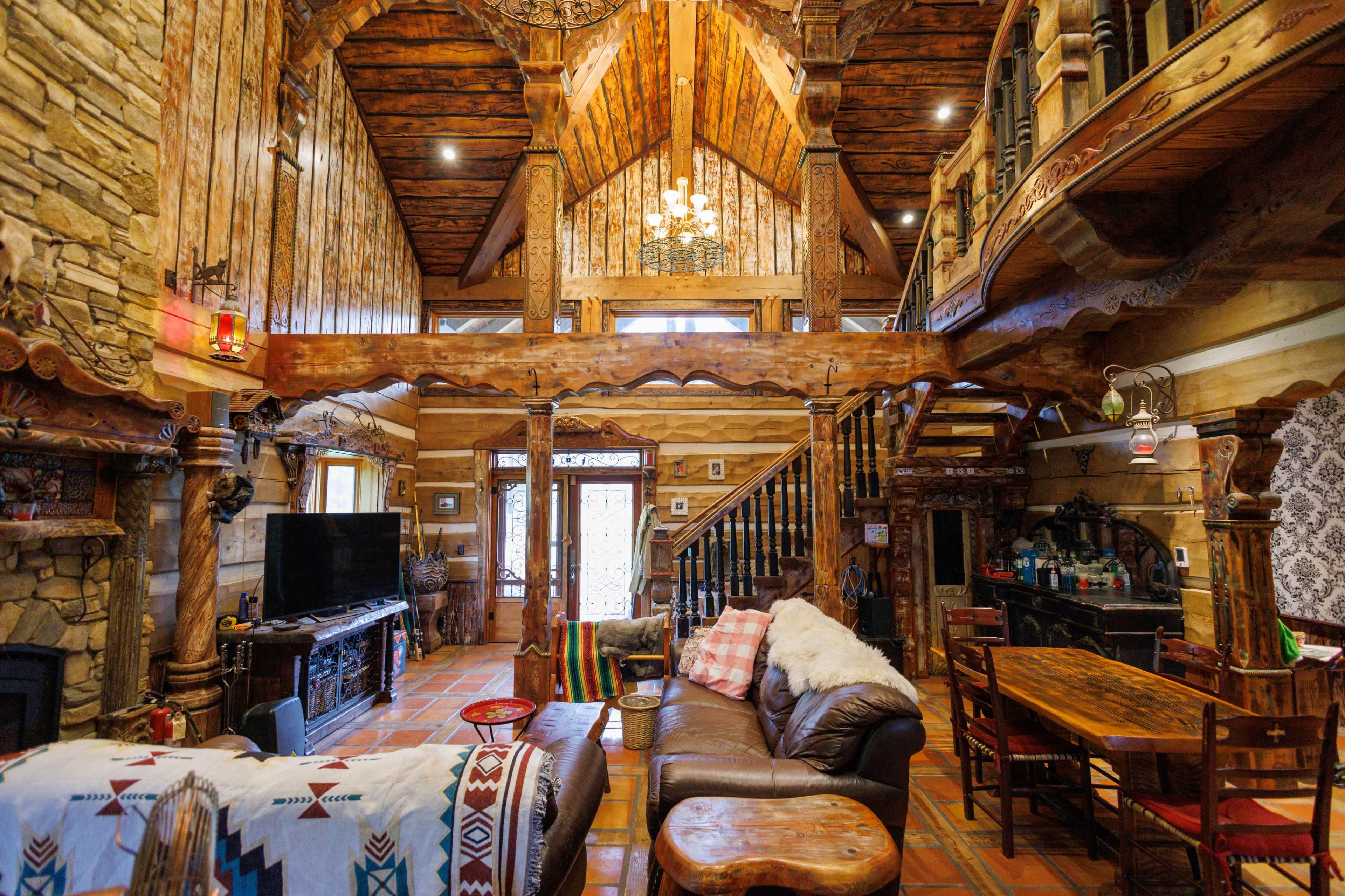 The image depicts a cozy, rustic cabin interior with wooden beams, stone accents, and a central staircase leading to a loft area.