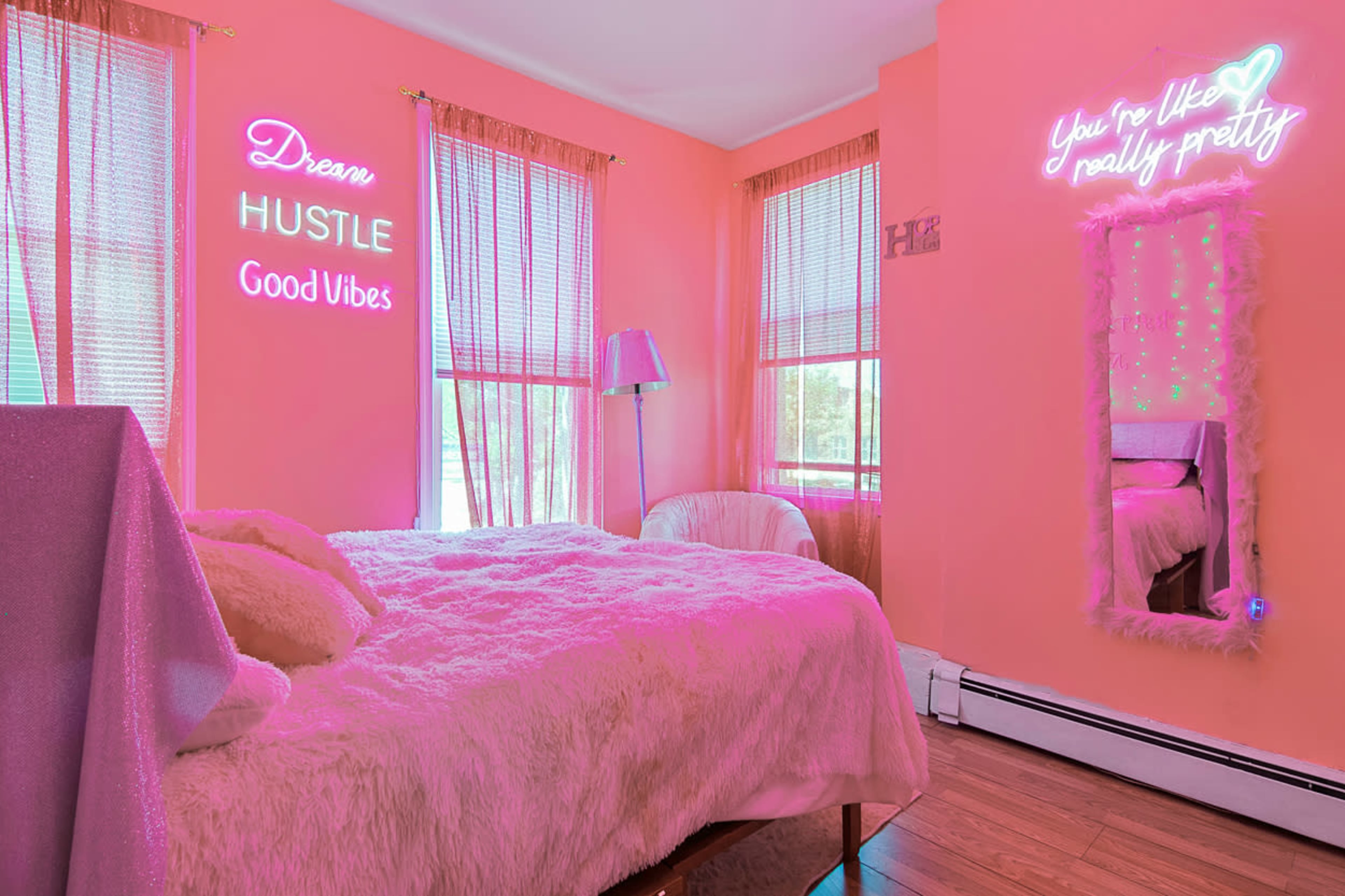 A brightly colored pink bedroom featuring a bed with a faux fur blanket, neon signs on the walls, and large windows letting in natural light.