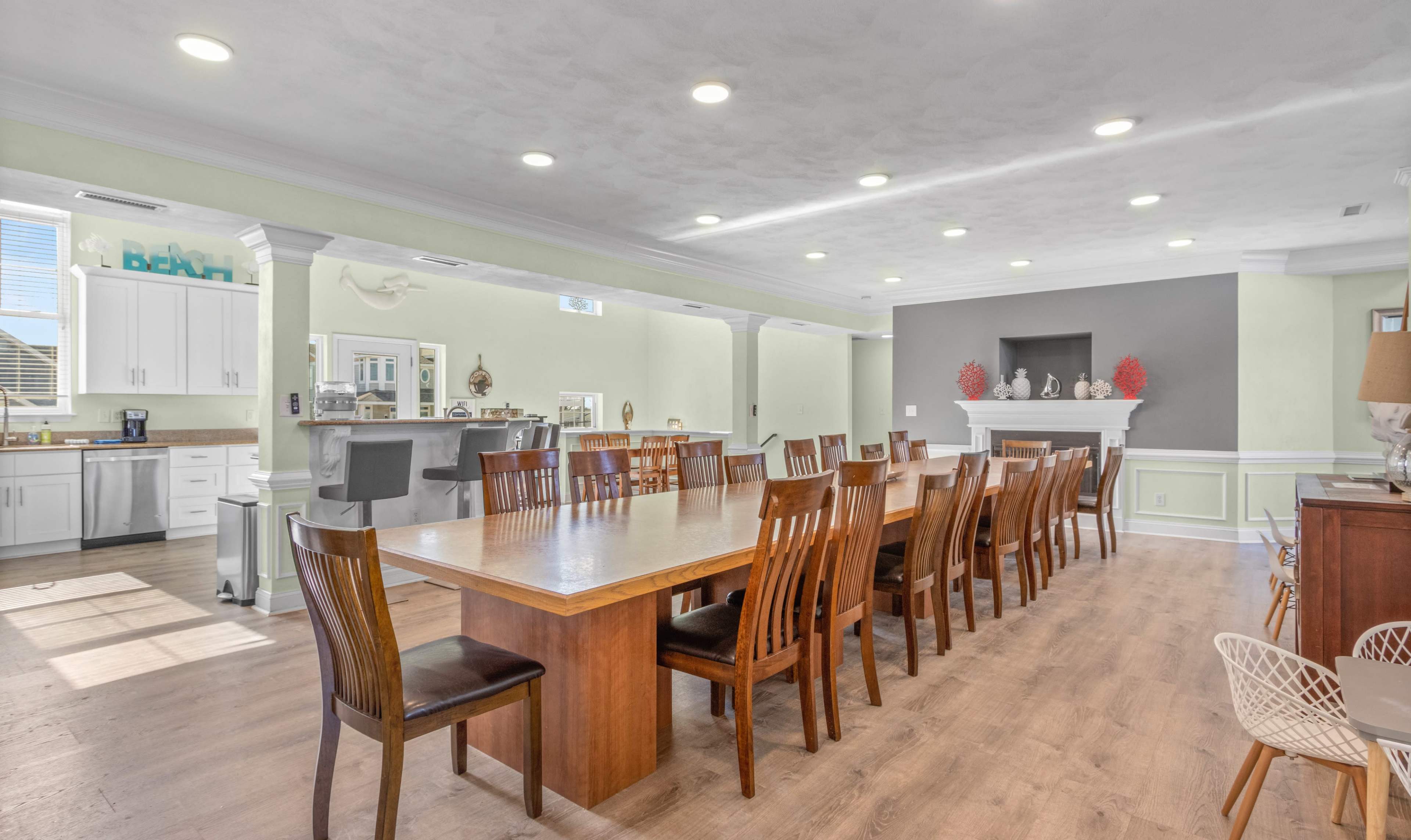 A long wooden dining table with multiple chairs is set in a spacious room that features a kitchen area and a fireplace.