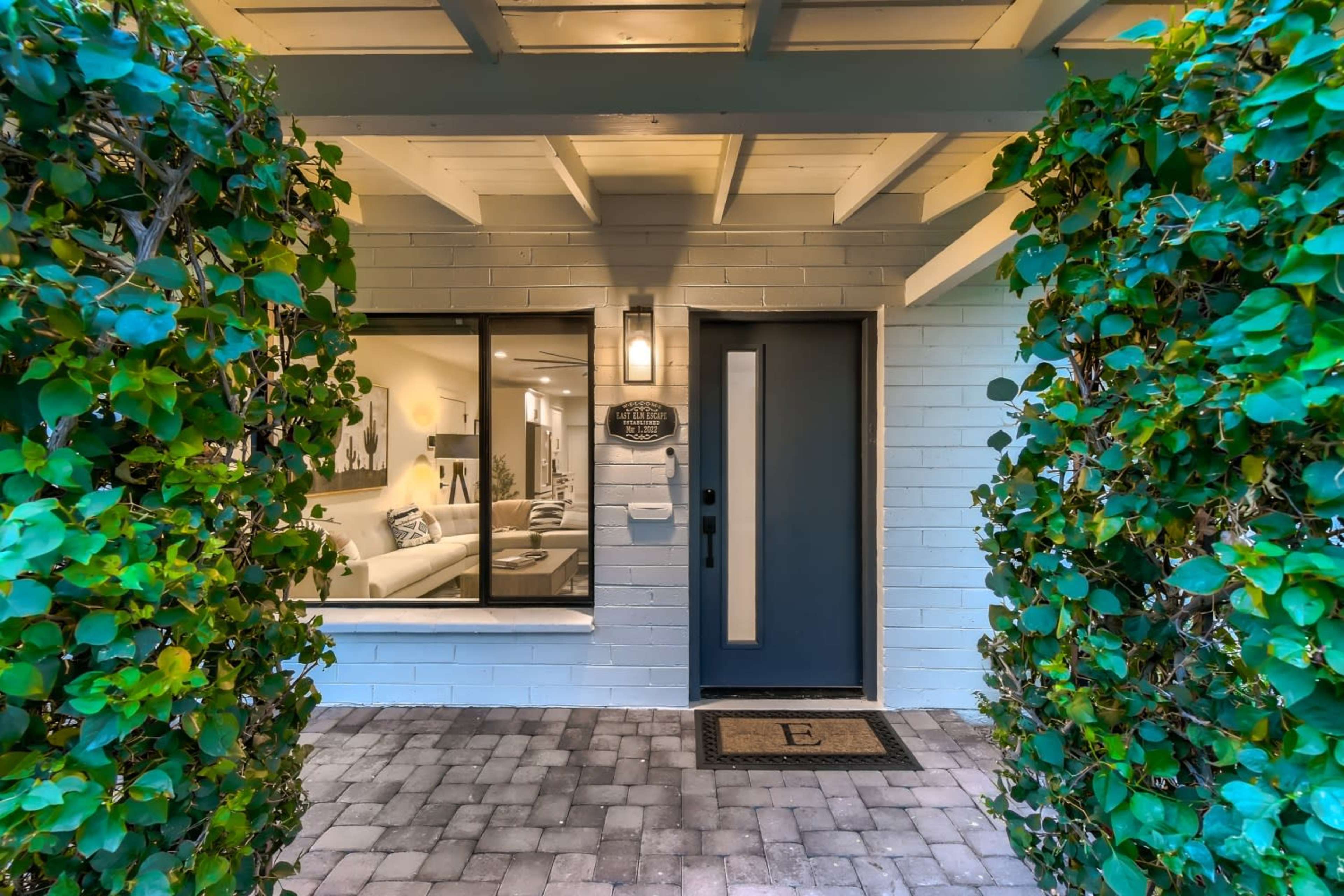 The entrance showcases a modern front door flanked by green foliage and a welcoming mat on a stone pathway.