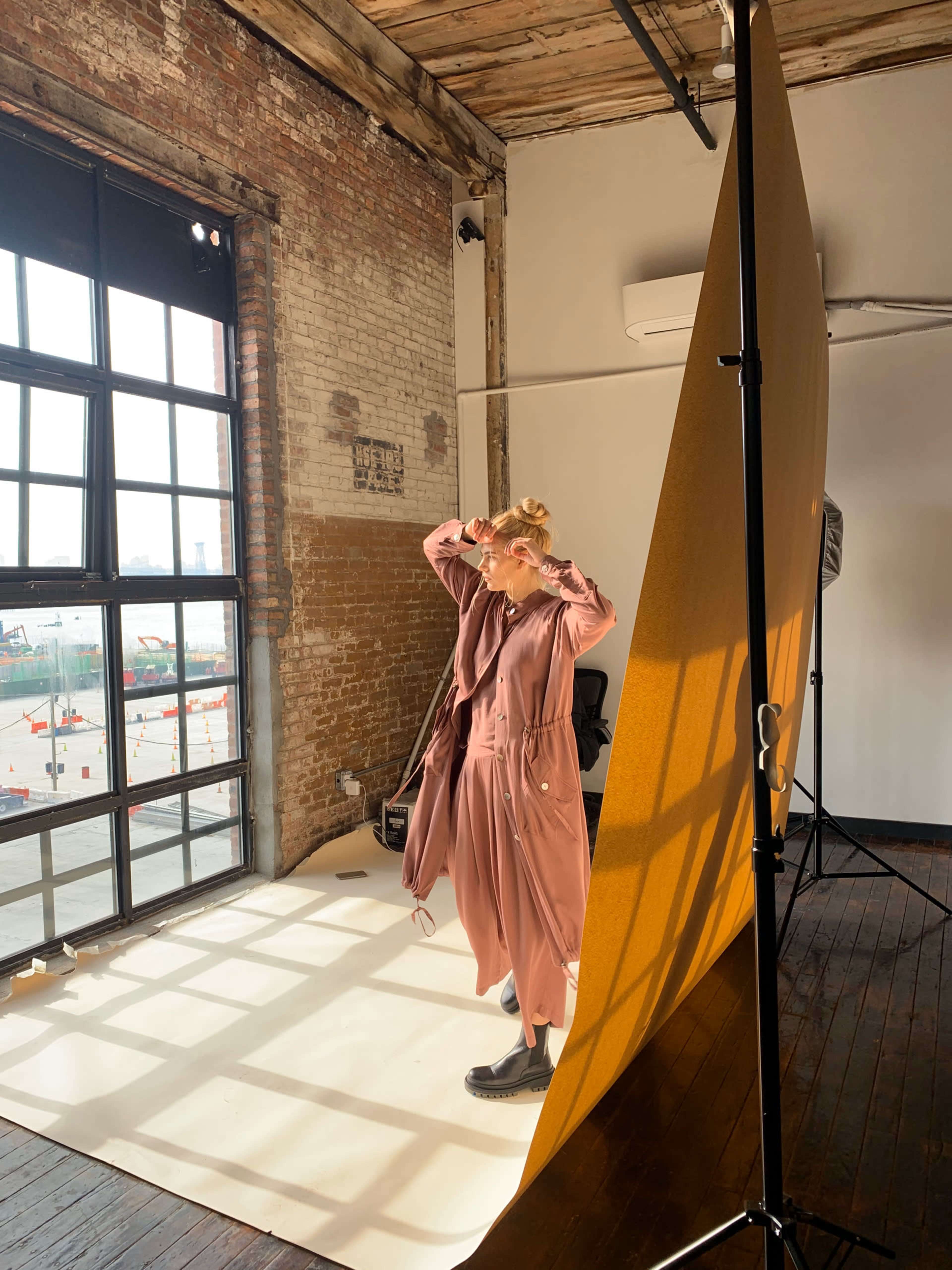 A person stands in front of a large window, posing against a backdrop while adjusting their hair in a well-lit studio space.