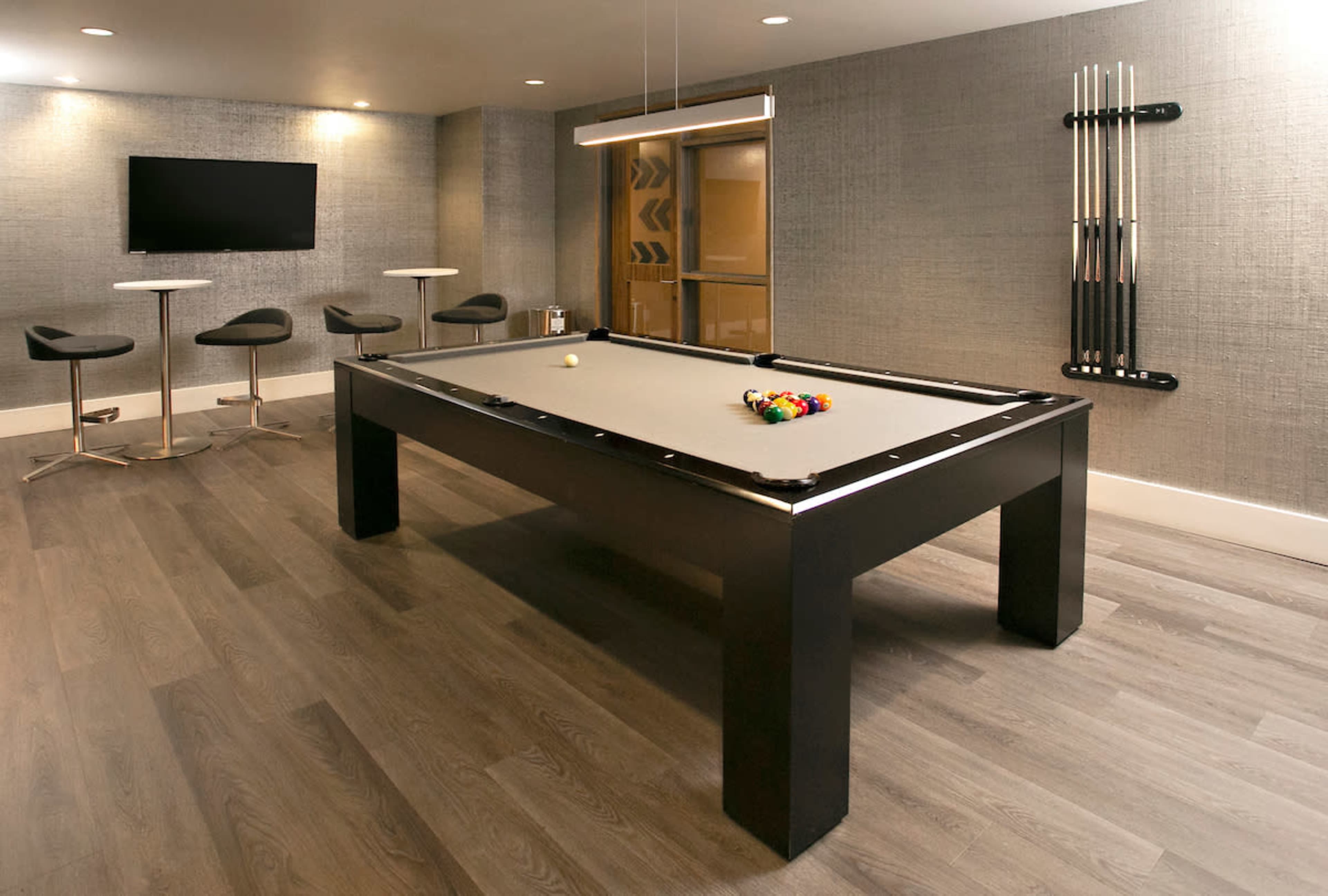A billiard table with a set of pool cues stands in a modern room featuring bar stools and a television on the wall.