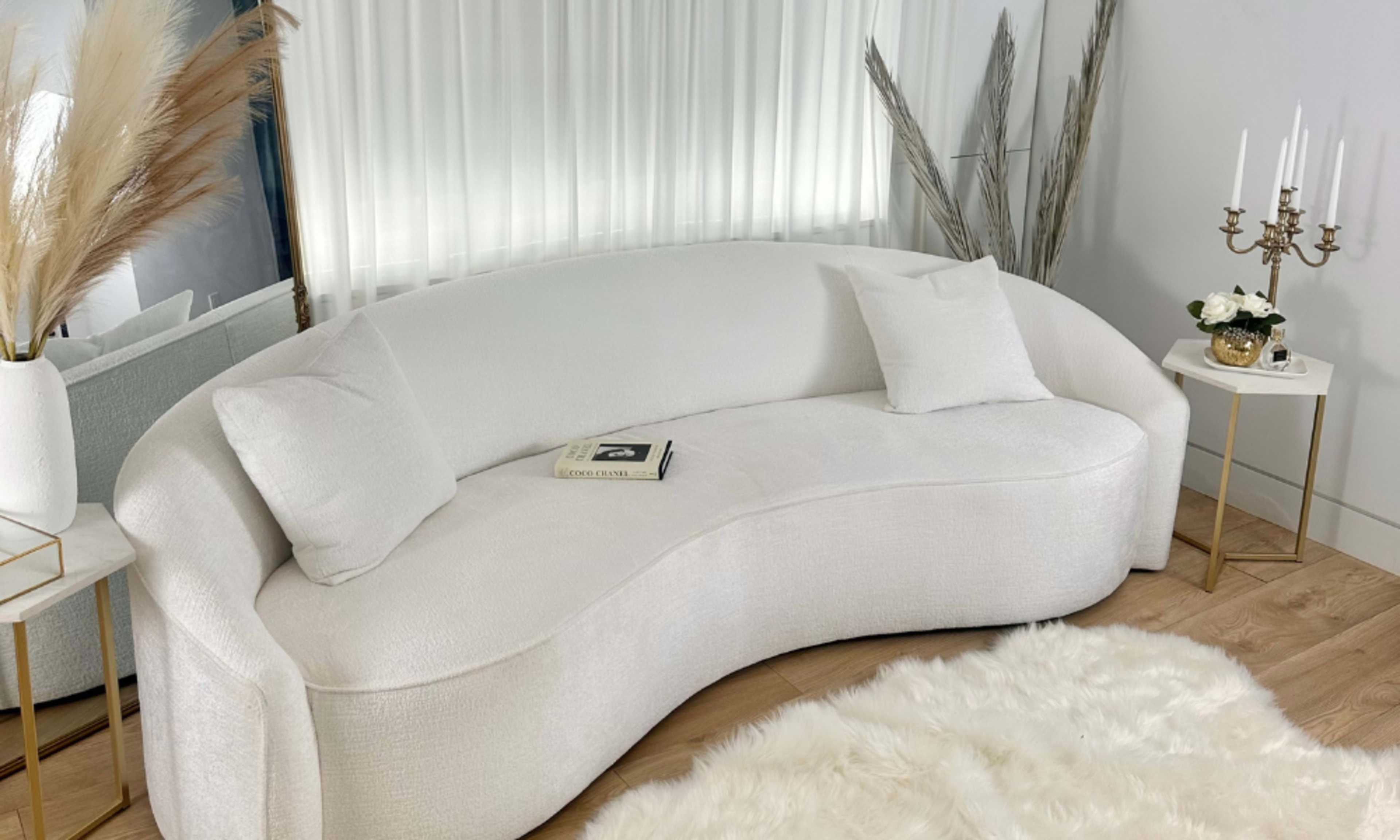 A curved white couch positioned in a well-lit room, accompanied by a small side table with candles and a decorative arrangement.