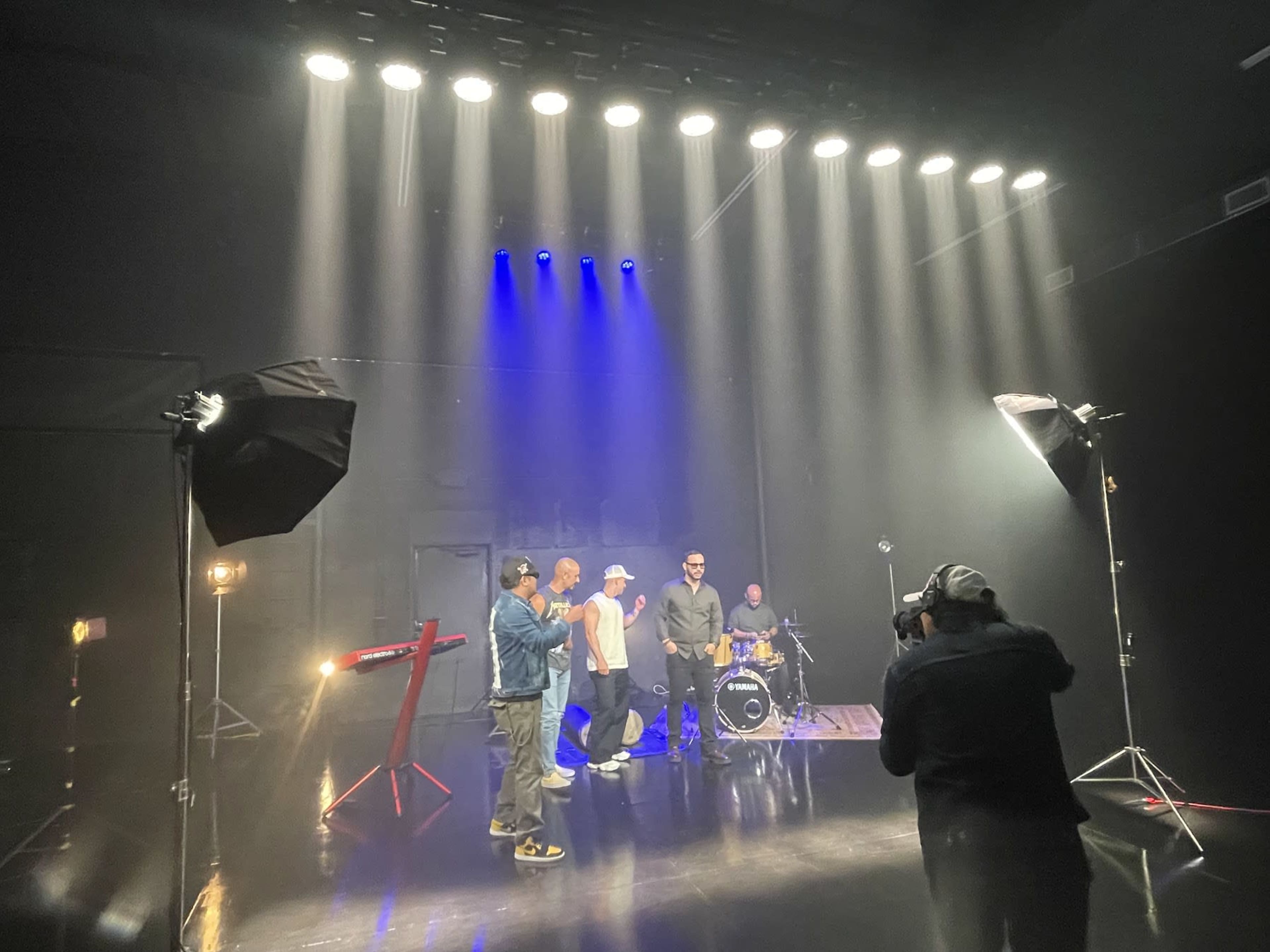 A band performs in a dimly lit studio with bright lights overhead and a photographer capturing the scene.