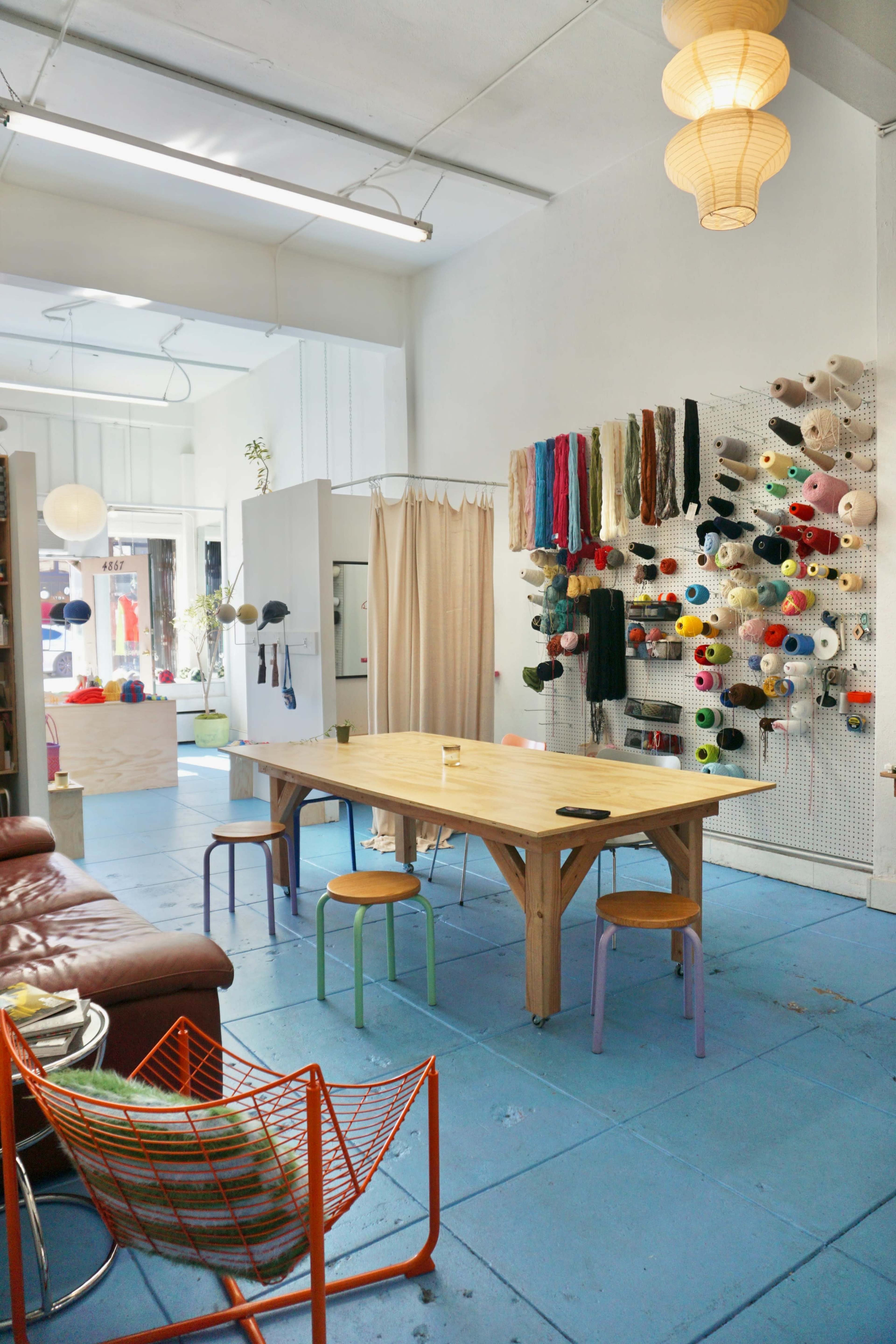The image shows a brightly lit craft space with a large wooden table surrounded by colorful chairs, and a wall displaying various spools of yarn.