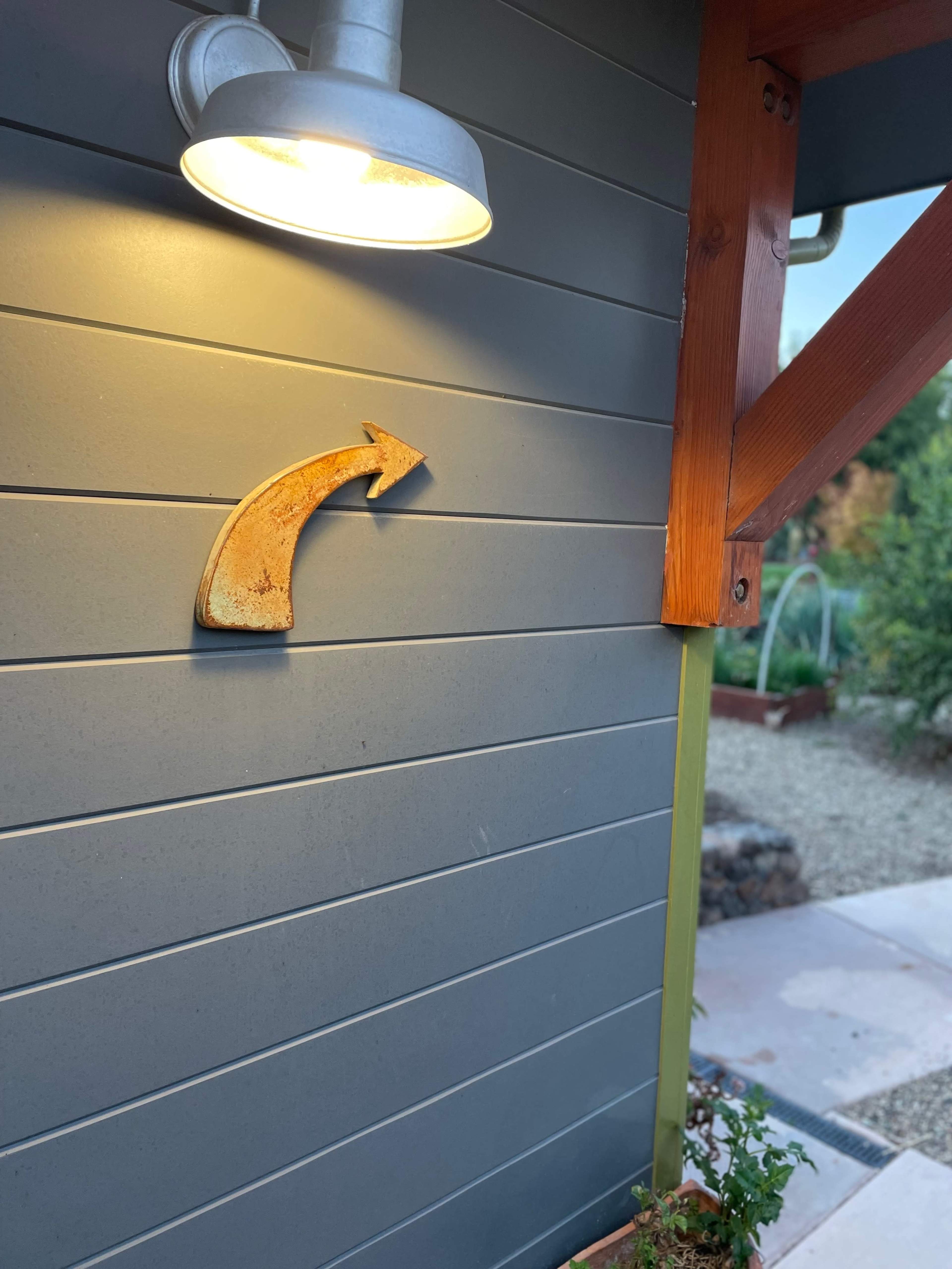 A weathered metal arrow sign points to the right on a gray wooden wall illuminated by a nearby light fixture.