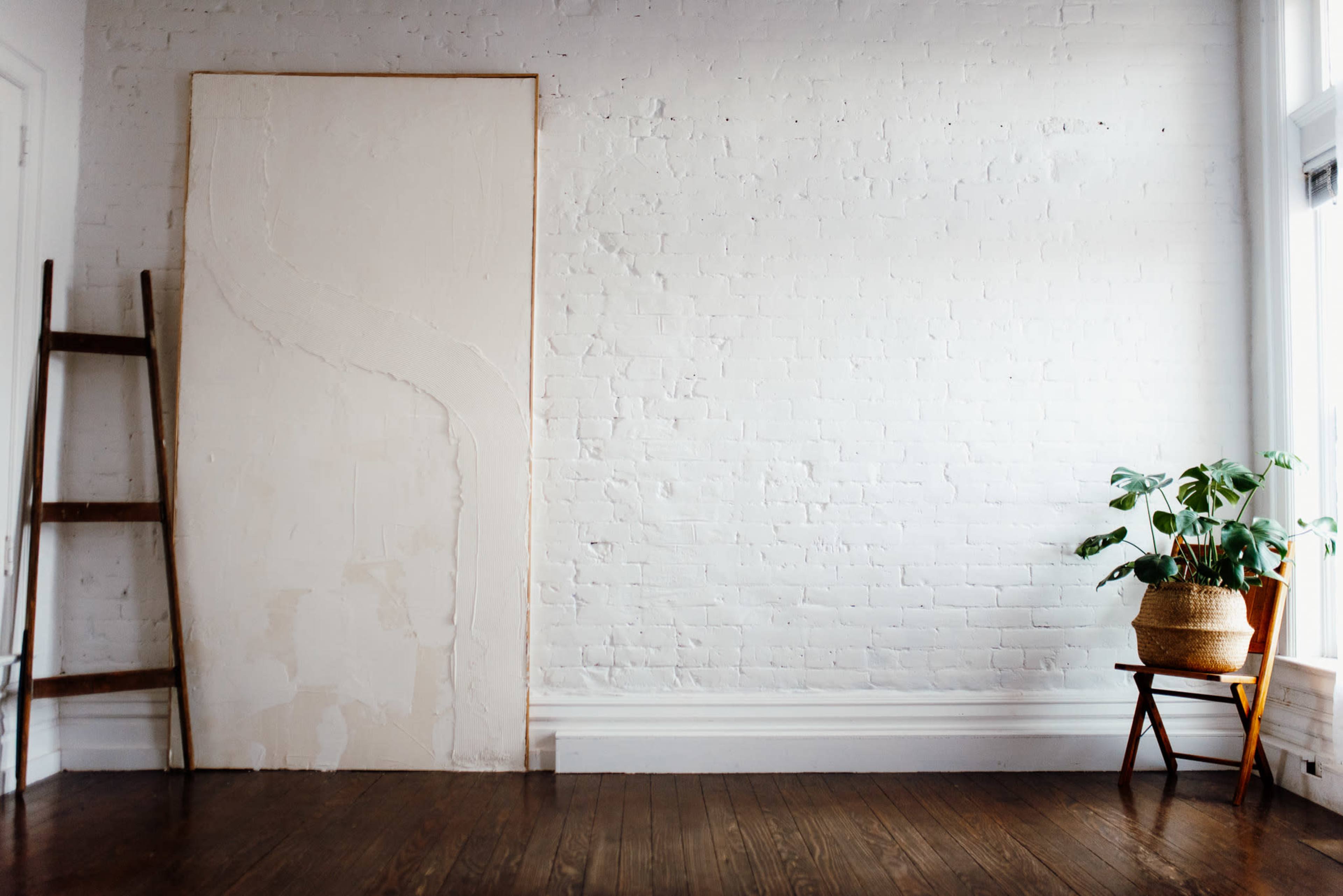 A wooden ladder leans against a white brick wall next to a large canvas and a chair with a potted plant on the floor beside it.