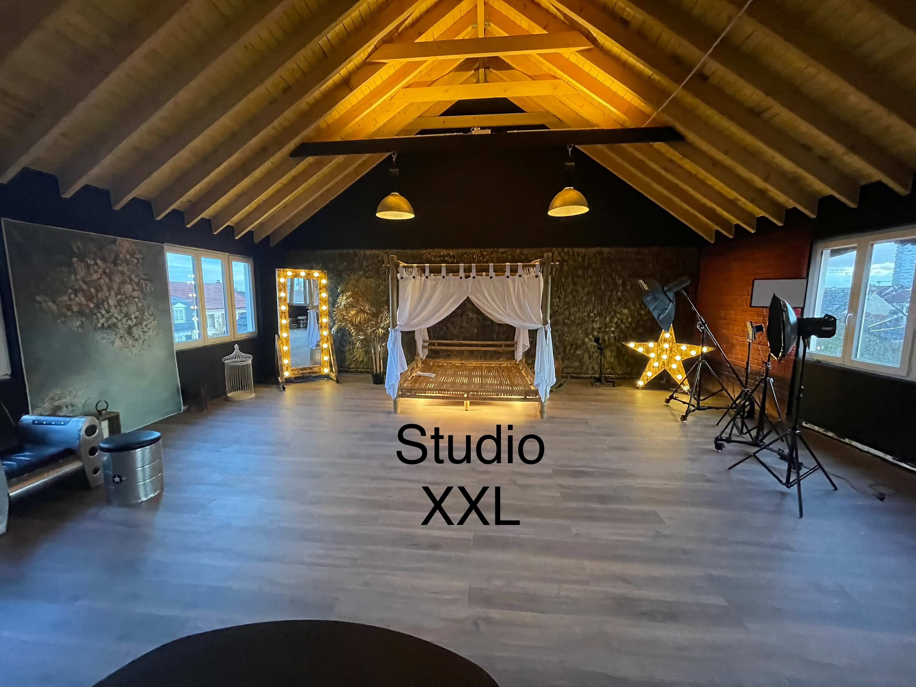 The image shows a spacious studio with wooden beams, featuring a decorated bed frame with curtains, illuminated letters, and photography equipment.