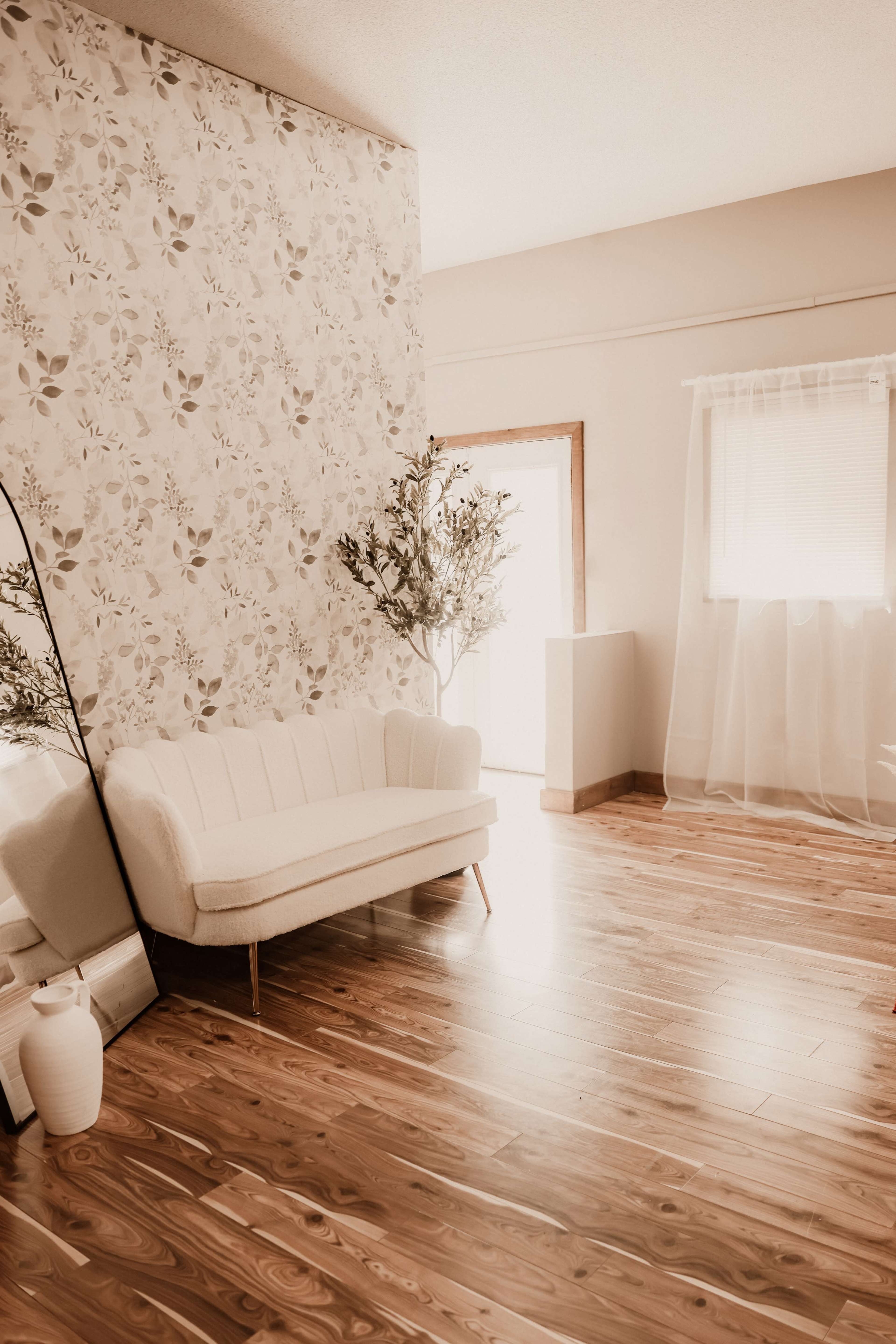 A bright, minimalist room with a floral wallpaper accent wall, a white sofa, a large mirror, a potted plant, and wooden flooring.