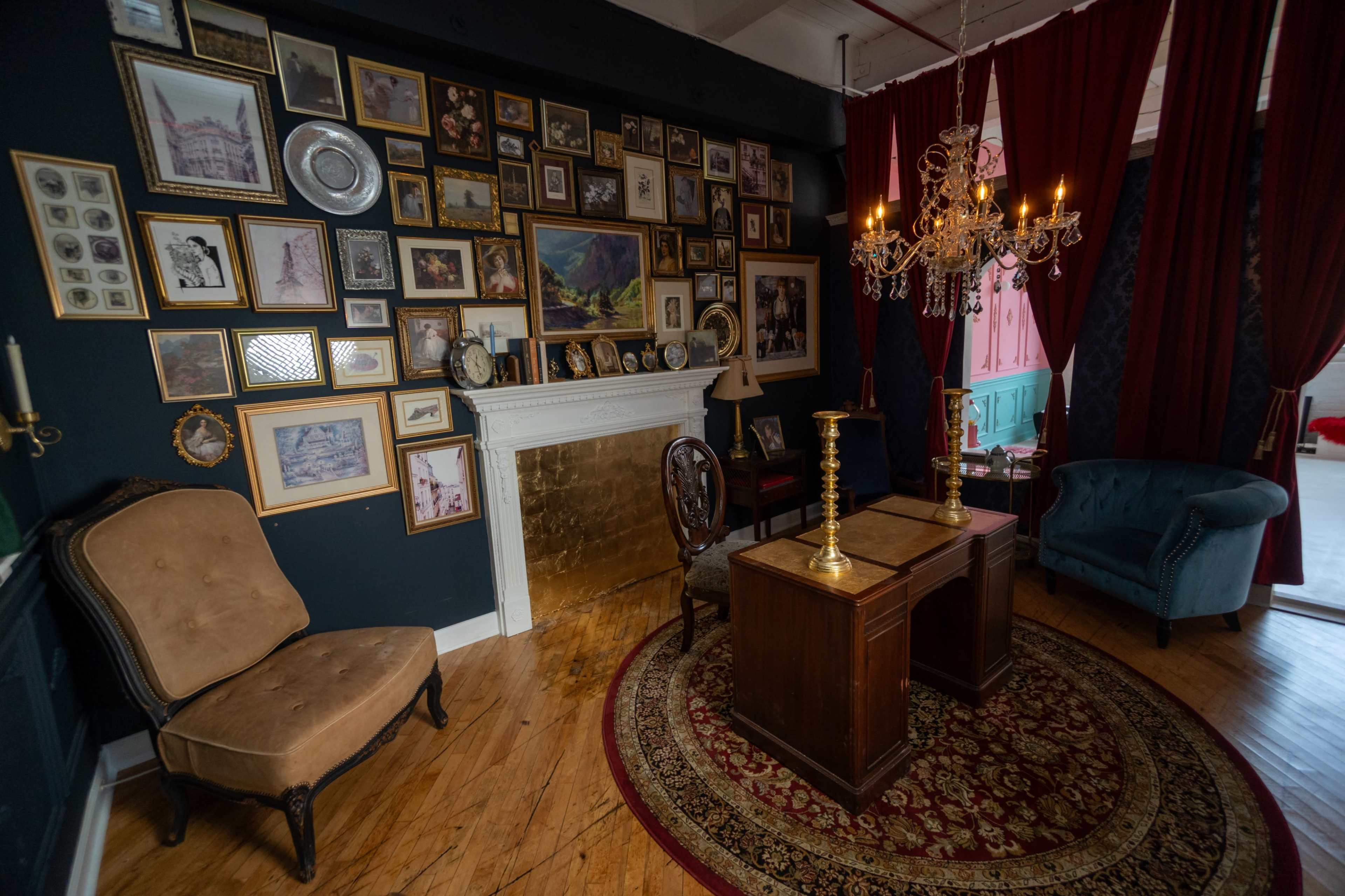 The image depicts a vintage-style room featuring a wall adorned with framed artwork and photographs, a decorative fireplace, a round rug, and two antique chairs positioned around a small wooden table.