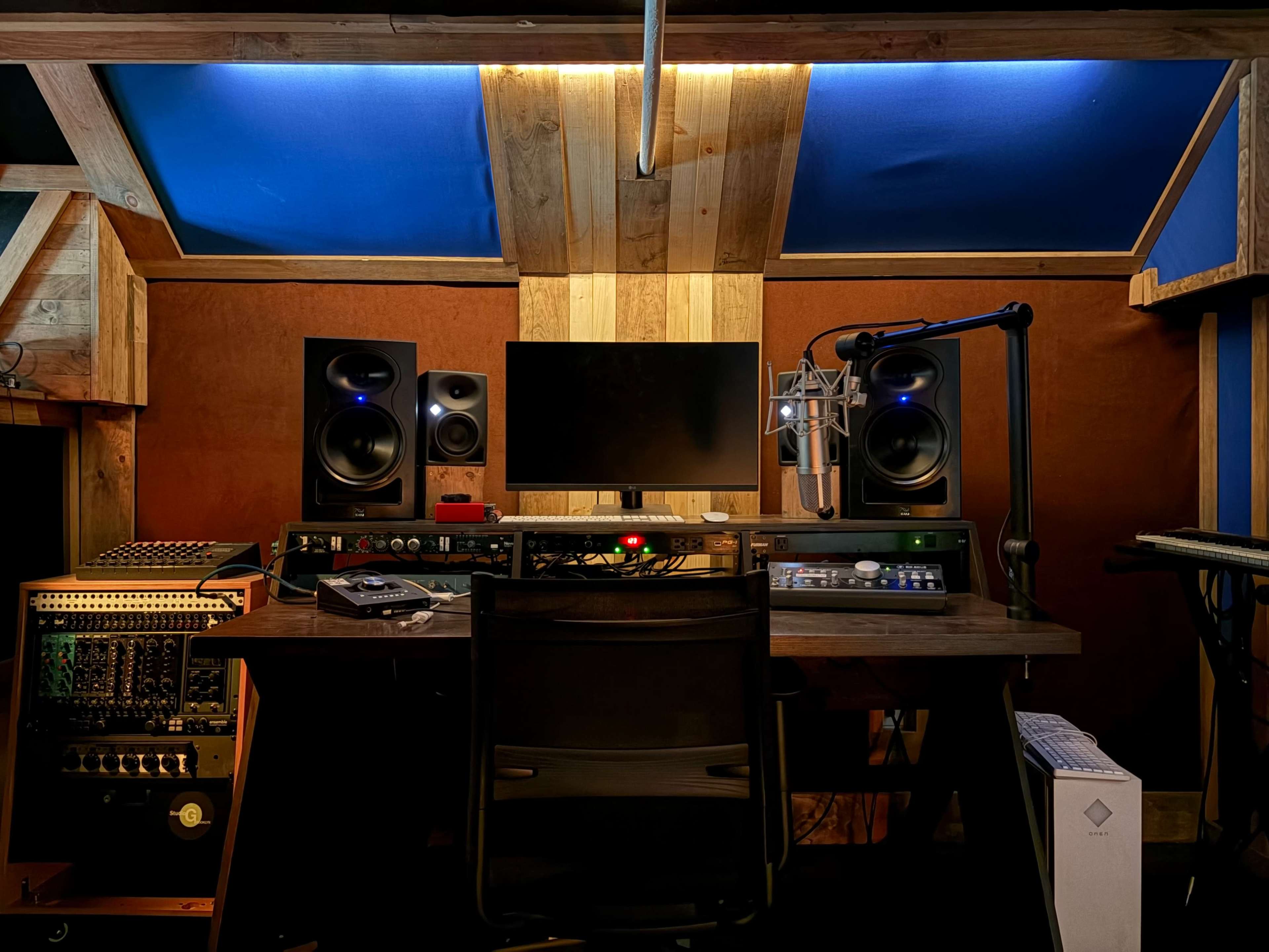 The image depicts a home recording studio with a desk set up for music production, featuring speakers, a computer monitor, a microphone, and various audio equipment.