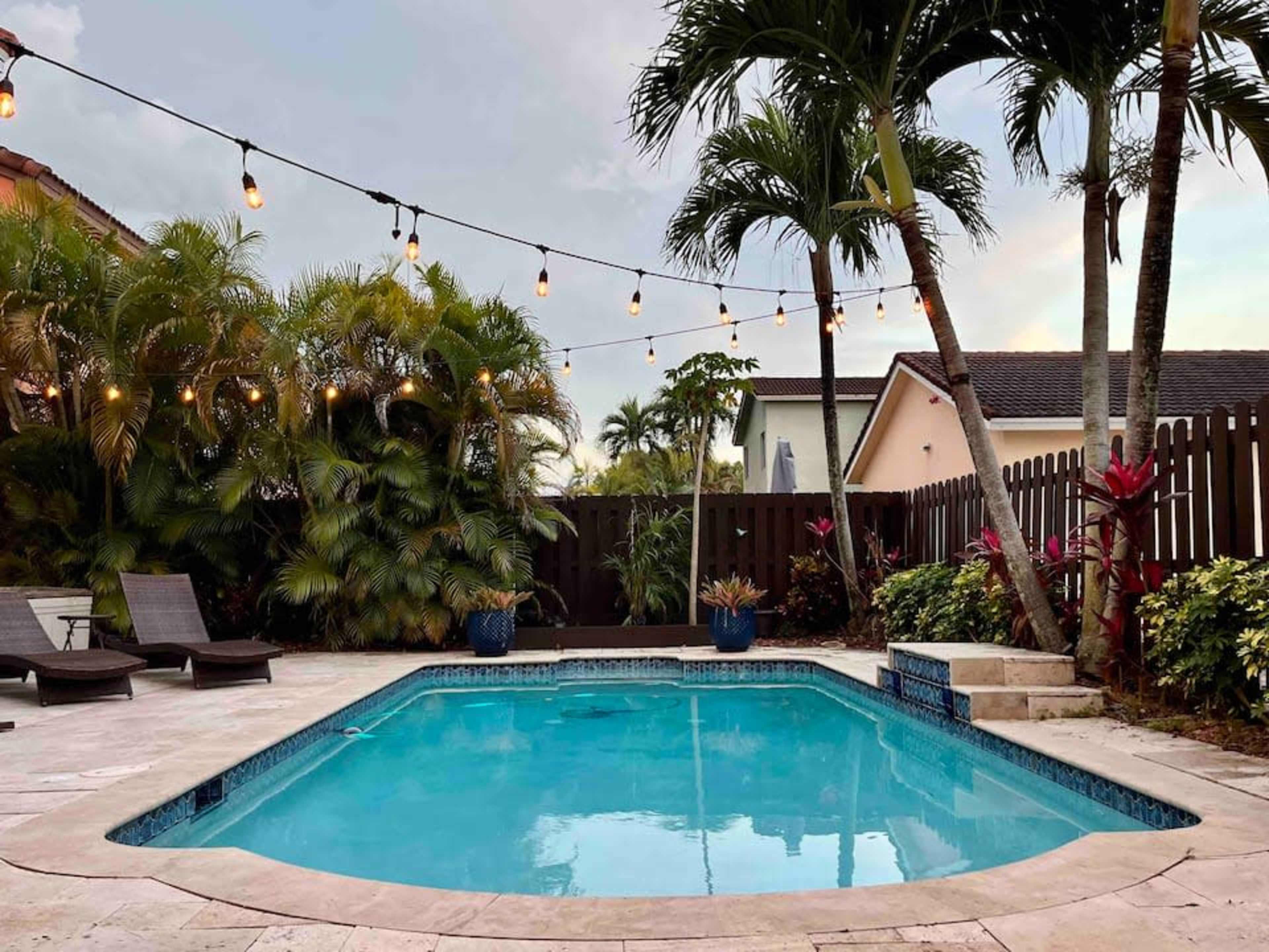A rectangular swimming pool is surrounded by palm trees and string lights, with lounge chairs nearby and tropical plants along the edges.
