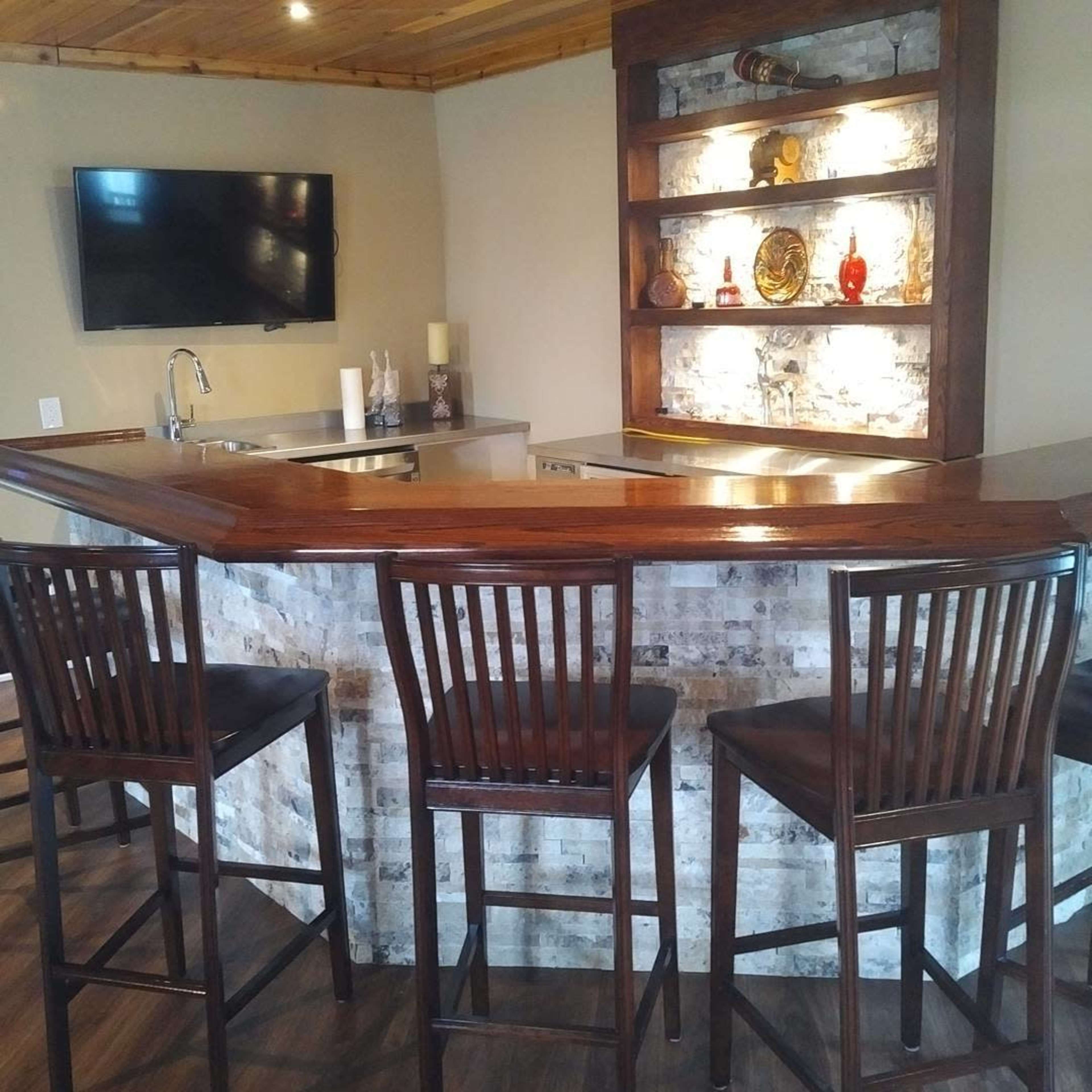 A modern home bar with a wooden counter, four high-backed chairs, and a display shelf with decorative items in a well-lit room.