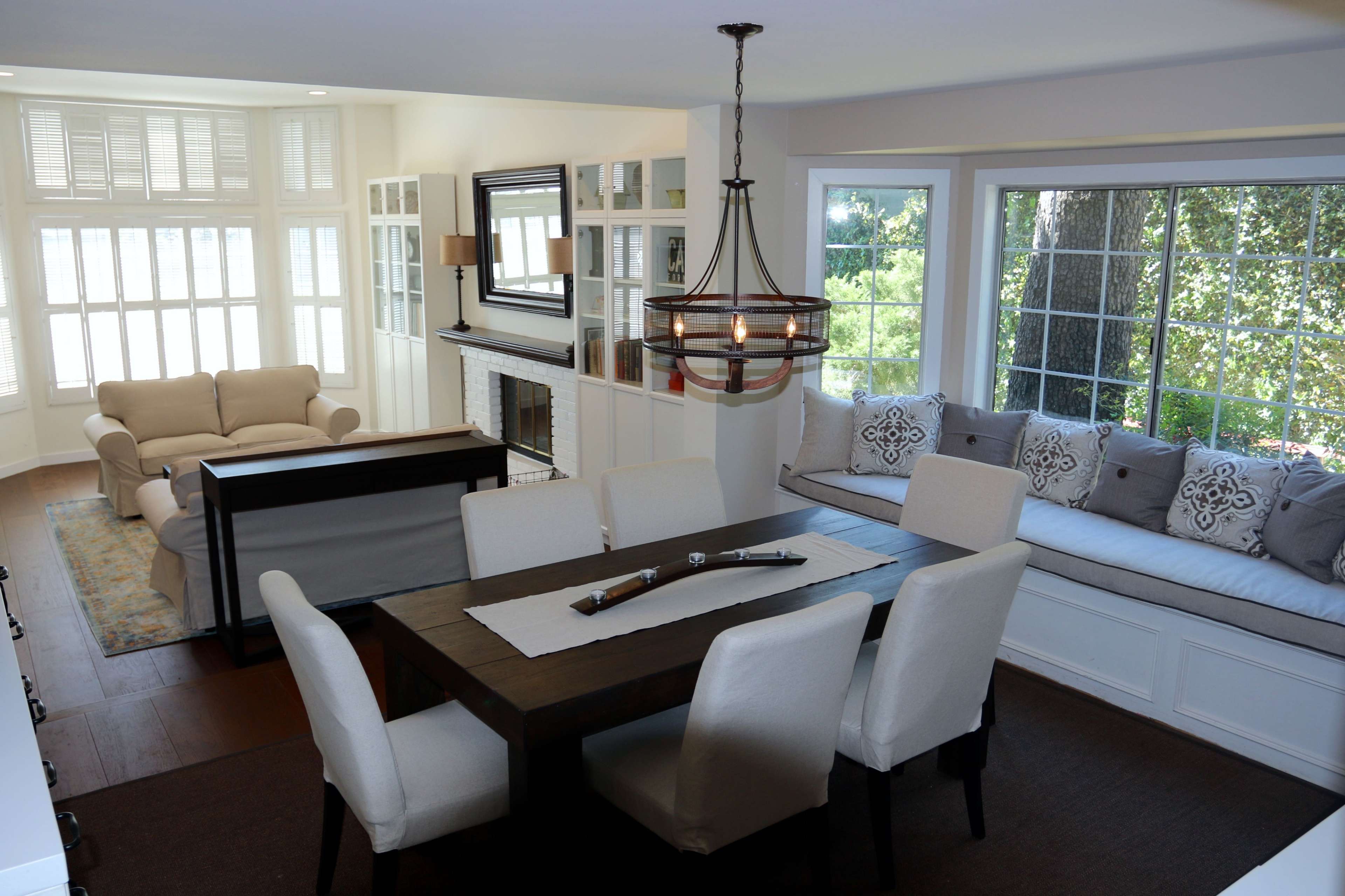 The image shows a modern, open-concept dining area with a wooden table surrounded by upholstered chairs, adjacent to a living space featuring a light-colored sofa and a built-in window seat.