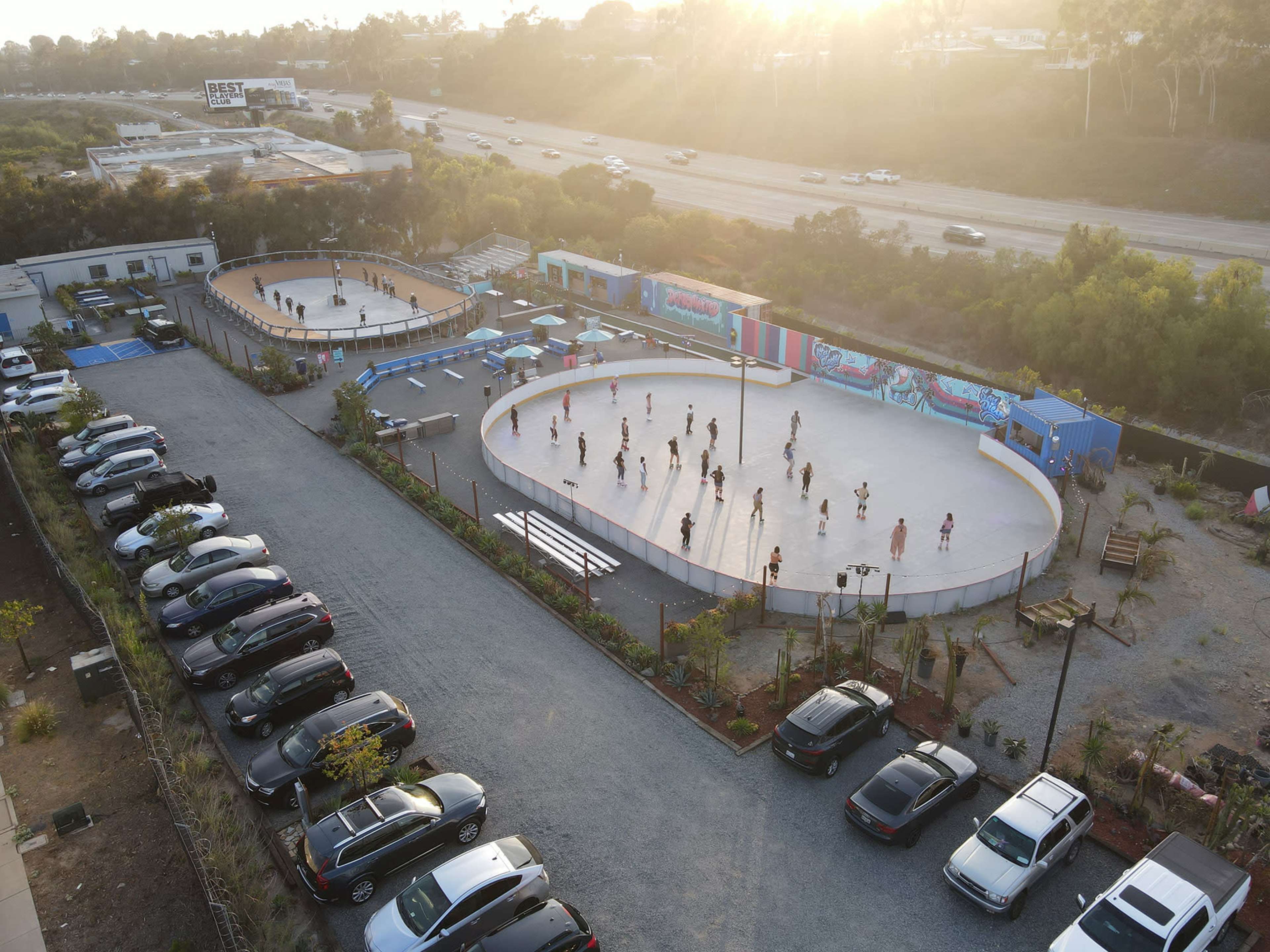 Two outdoor skating rinks with people skating, surrounded by cars parked in a gravel lot and bordered by greenery, under a sunset.