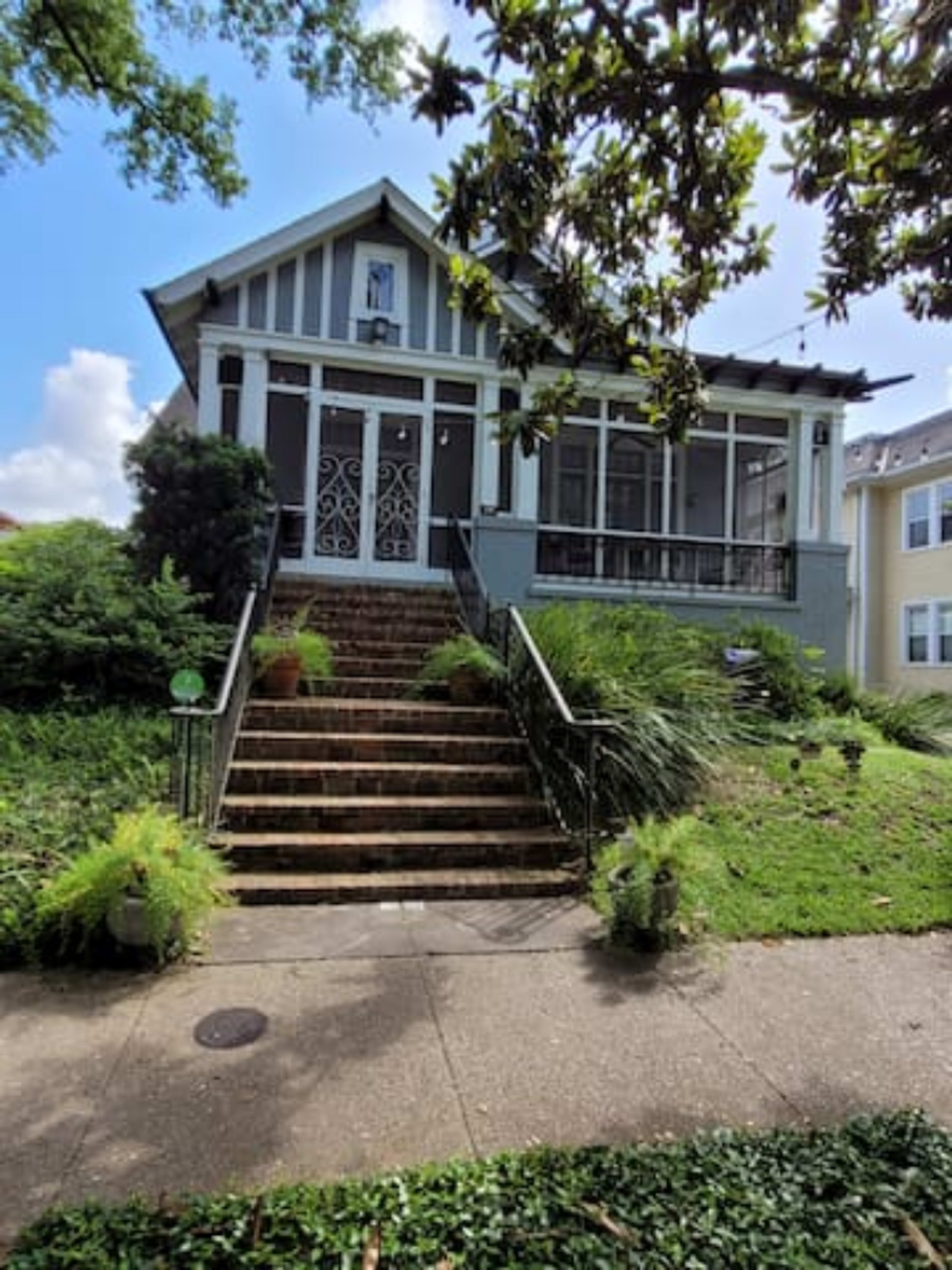 A two-story house with a front porch and decorative railings is set on a grassy yard with stone steps leading to the entrance.