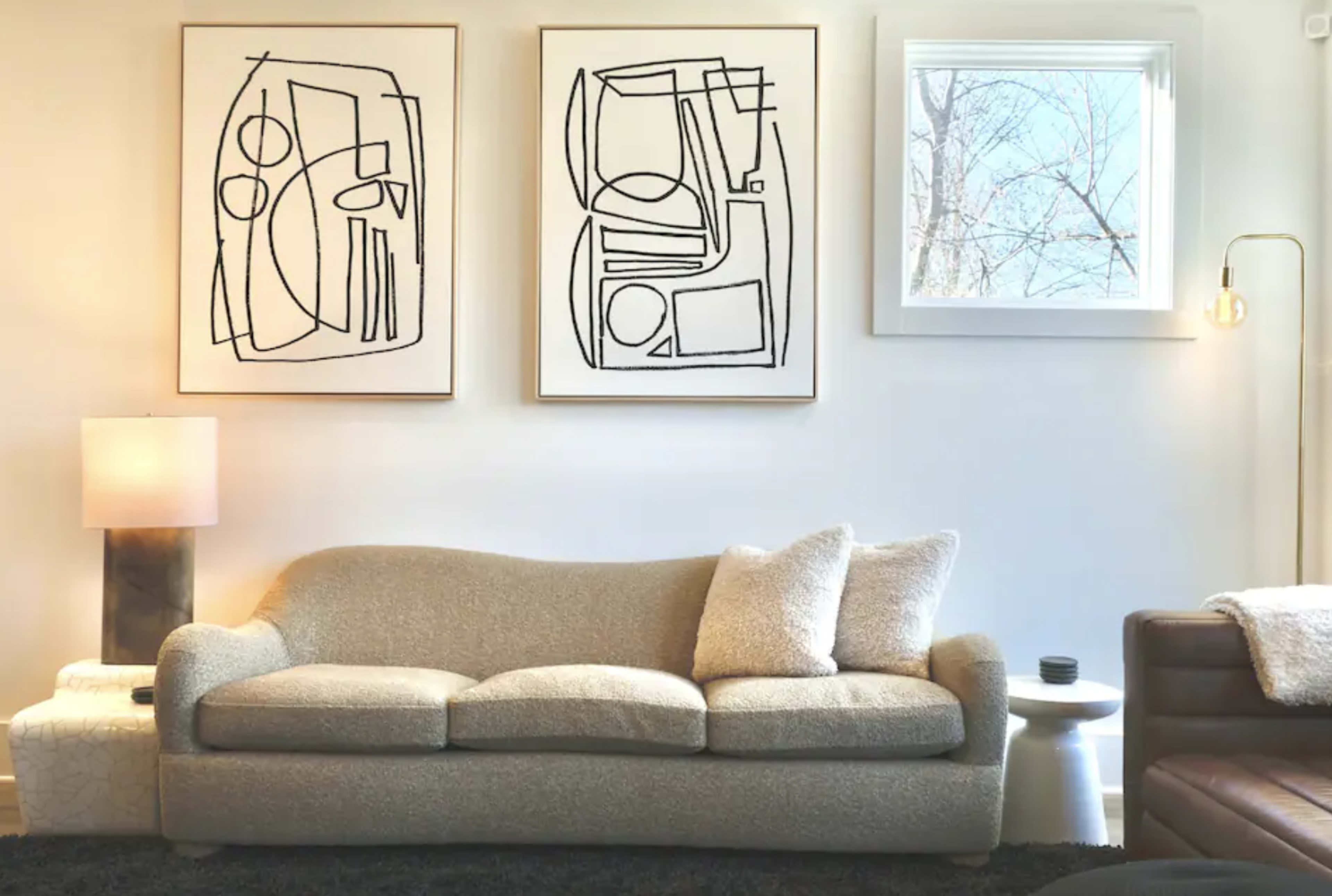 A living room features a light-colored sofa with textured upholstery, two abstract line art pieces on the wall, and a window allowing natural light to enter.