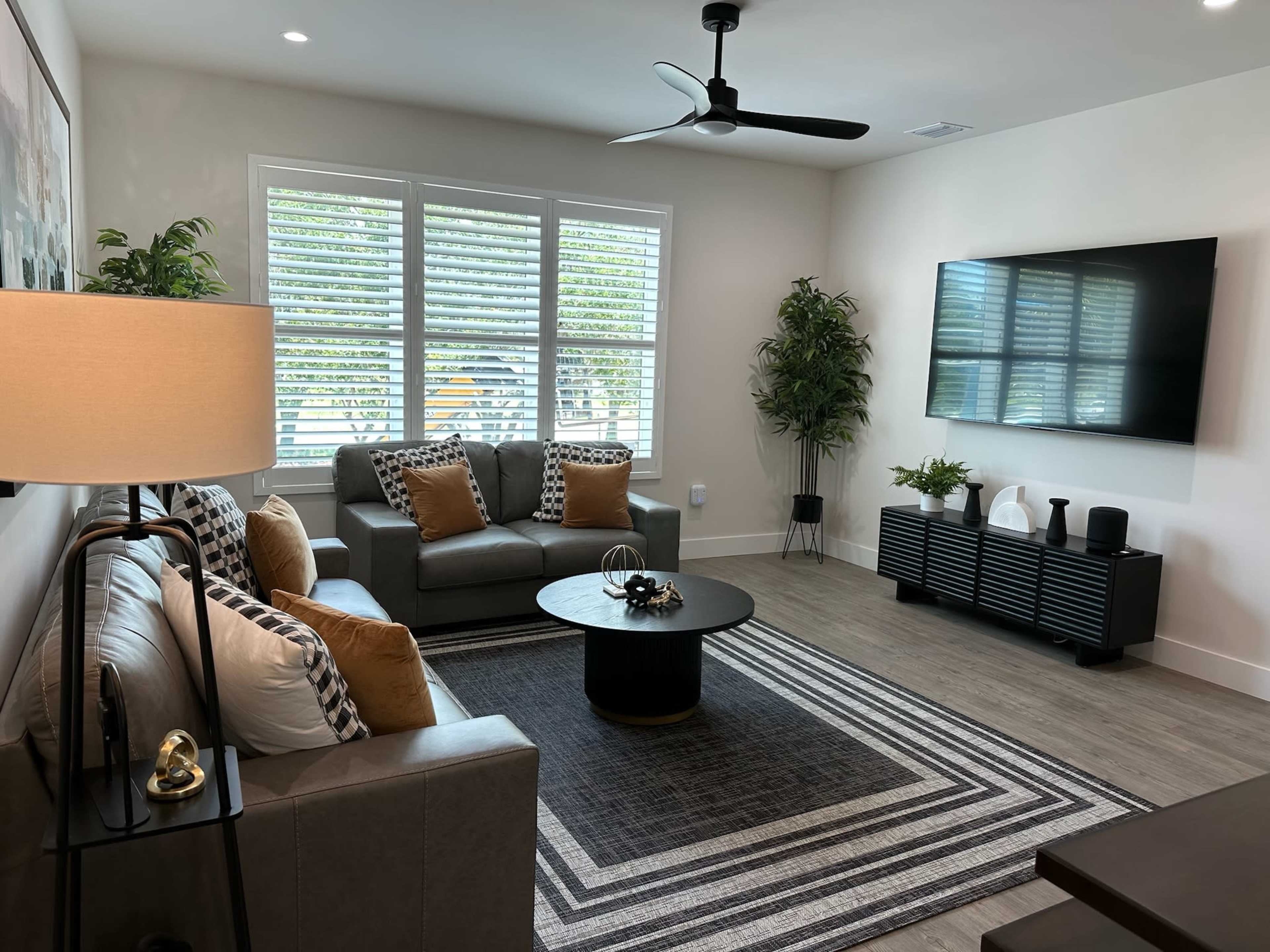 A modern living room featuring a gray sectional sofa, a round black coffee table, and a large wall-mounted TV, complemented by natural light streaming through slatted windows.