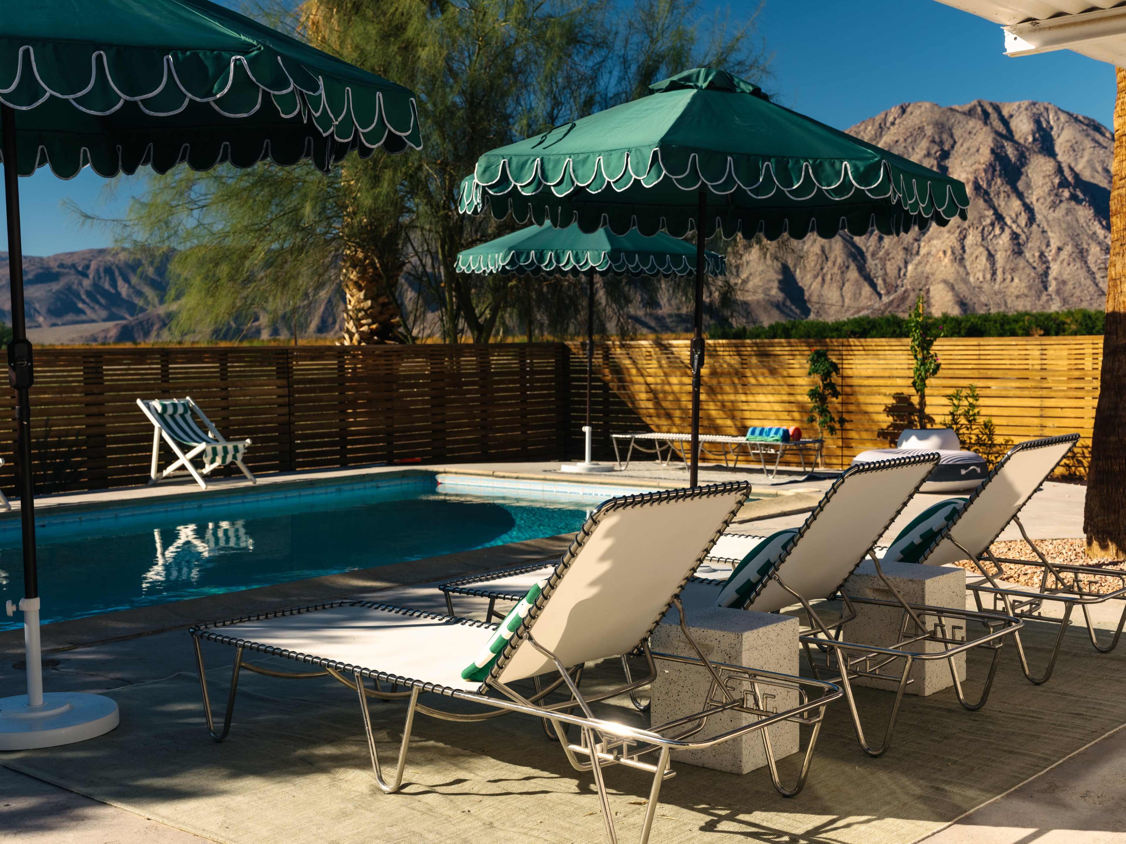 Lounge chairs with green umbrellas surround a swimming pool in a desert landscape with mountains in the background.