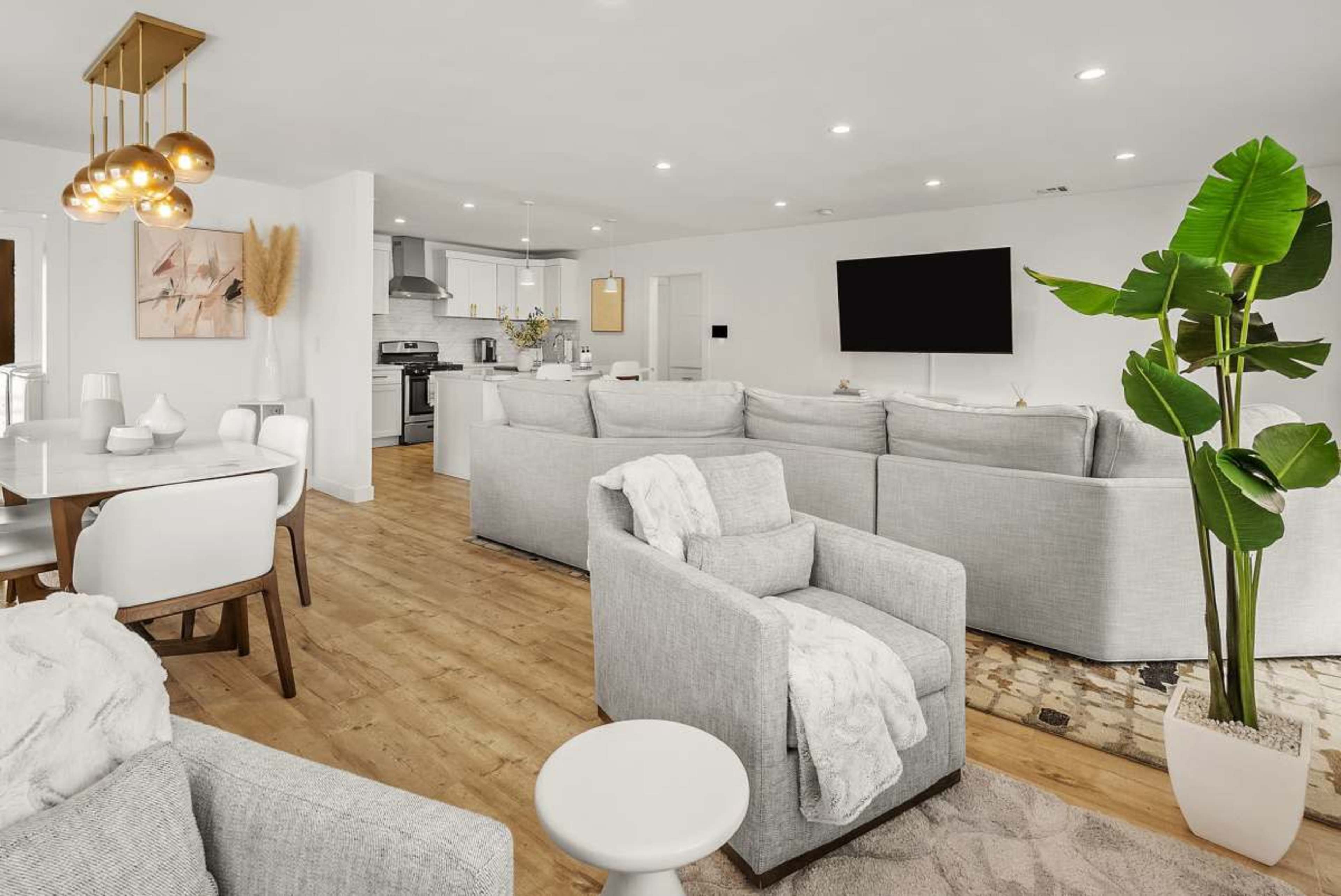 The image depicts a modern open-concept living and dining area featuring light gray furniture, a dining table, and a large television mounted on the wall.