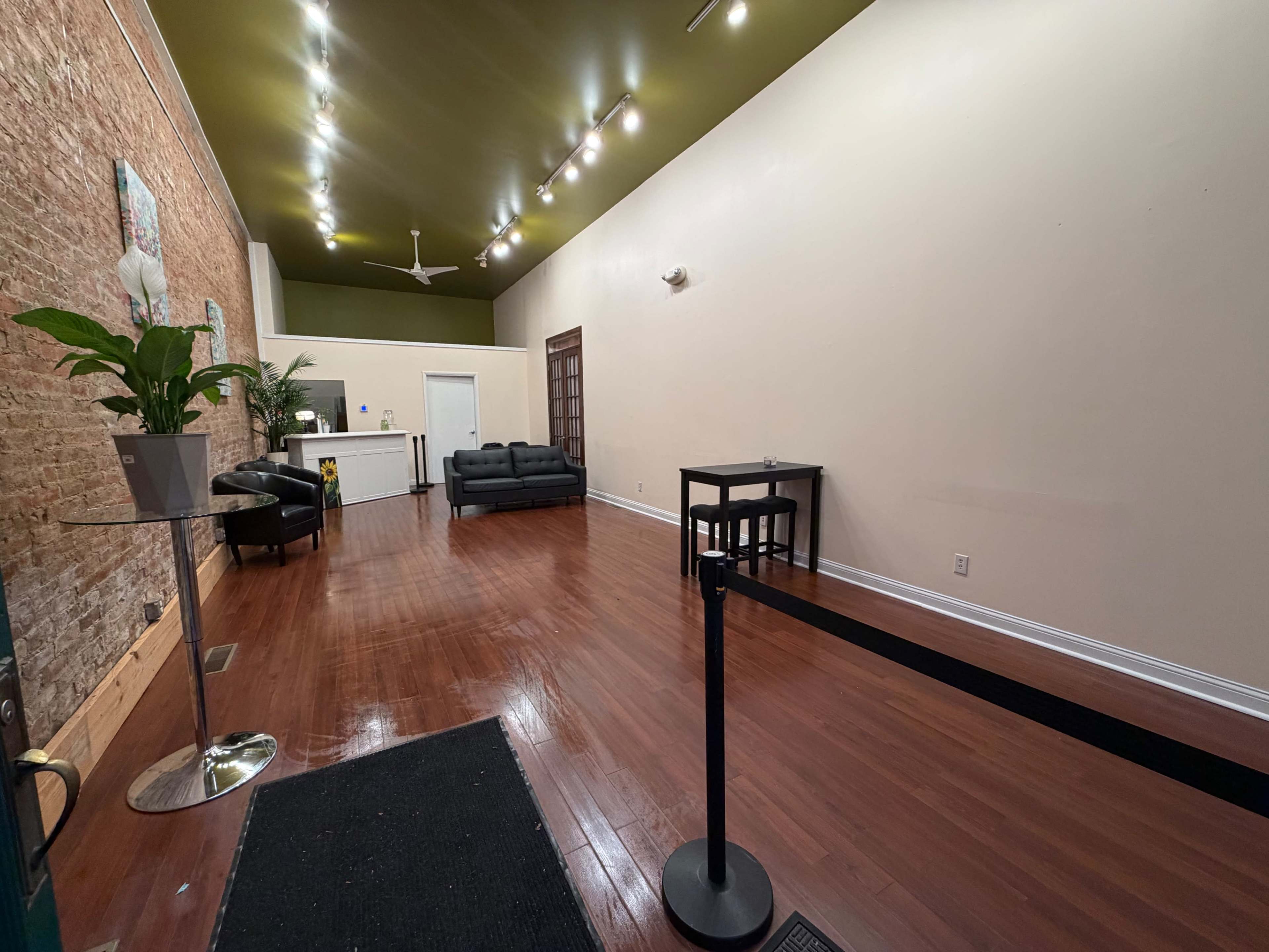 The image shows a spacious, empty room with polished wooden floors, a reception area at the back, and minimal furniture including two sofas, a small table, and a waiting line barrier.