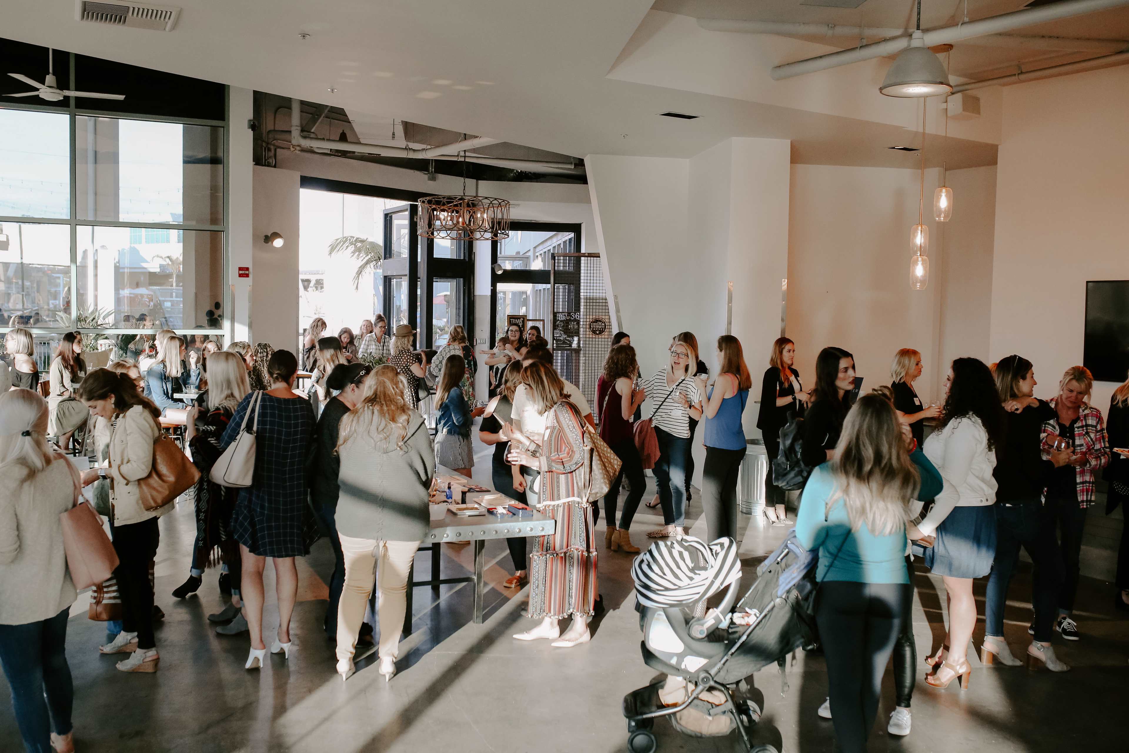 A large group of people gathers in a modern indoor space, engaging in conversation and browsing various tables.