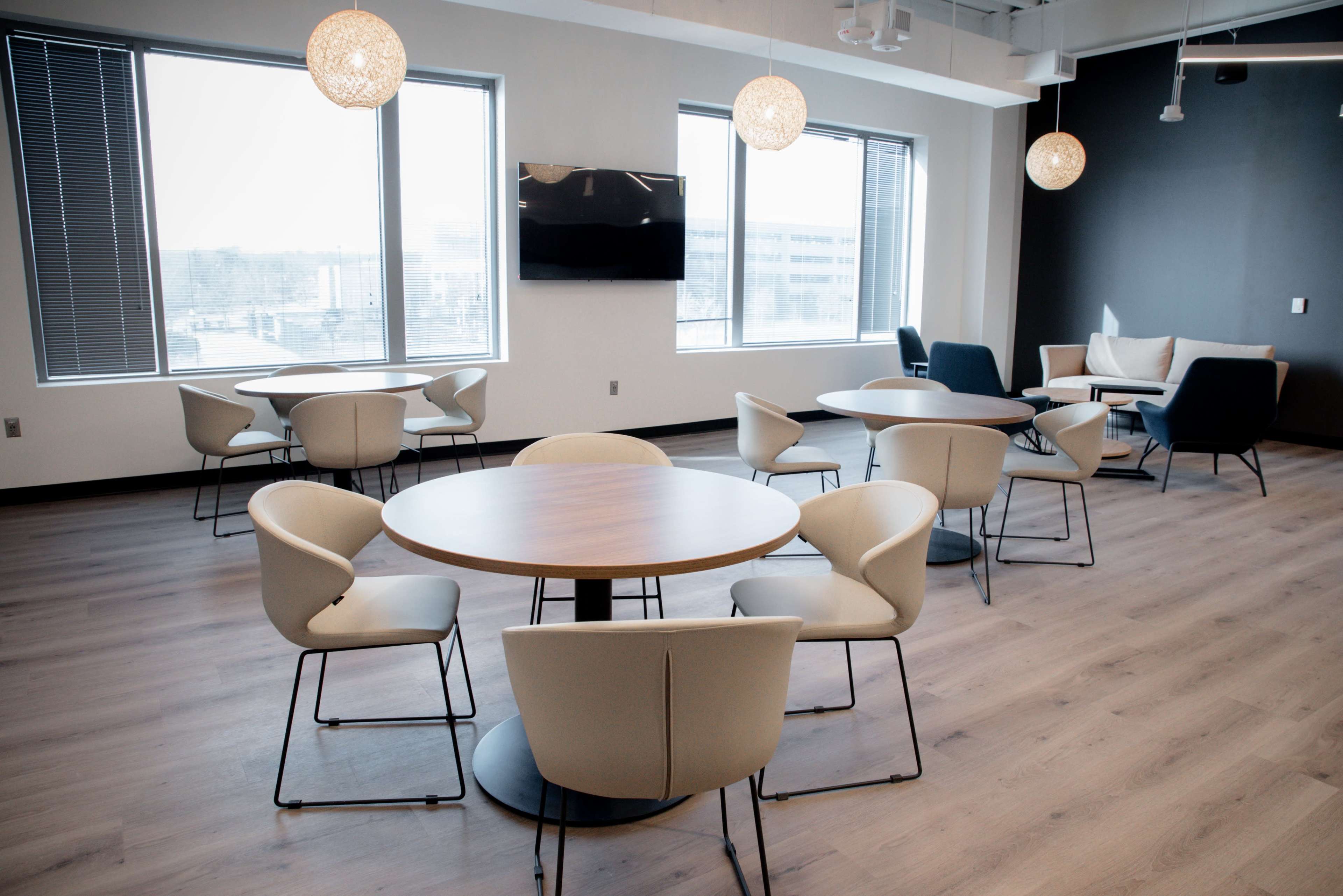 The image shows a modern office lounge area with several round tables and chairs, large windows, and pendant lights.