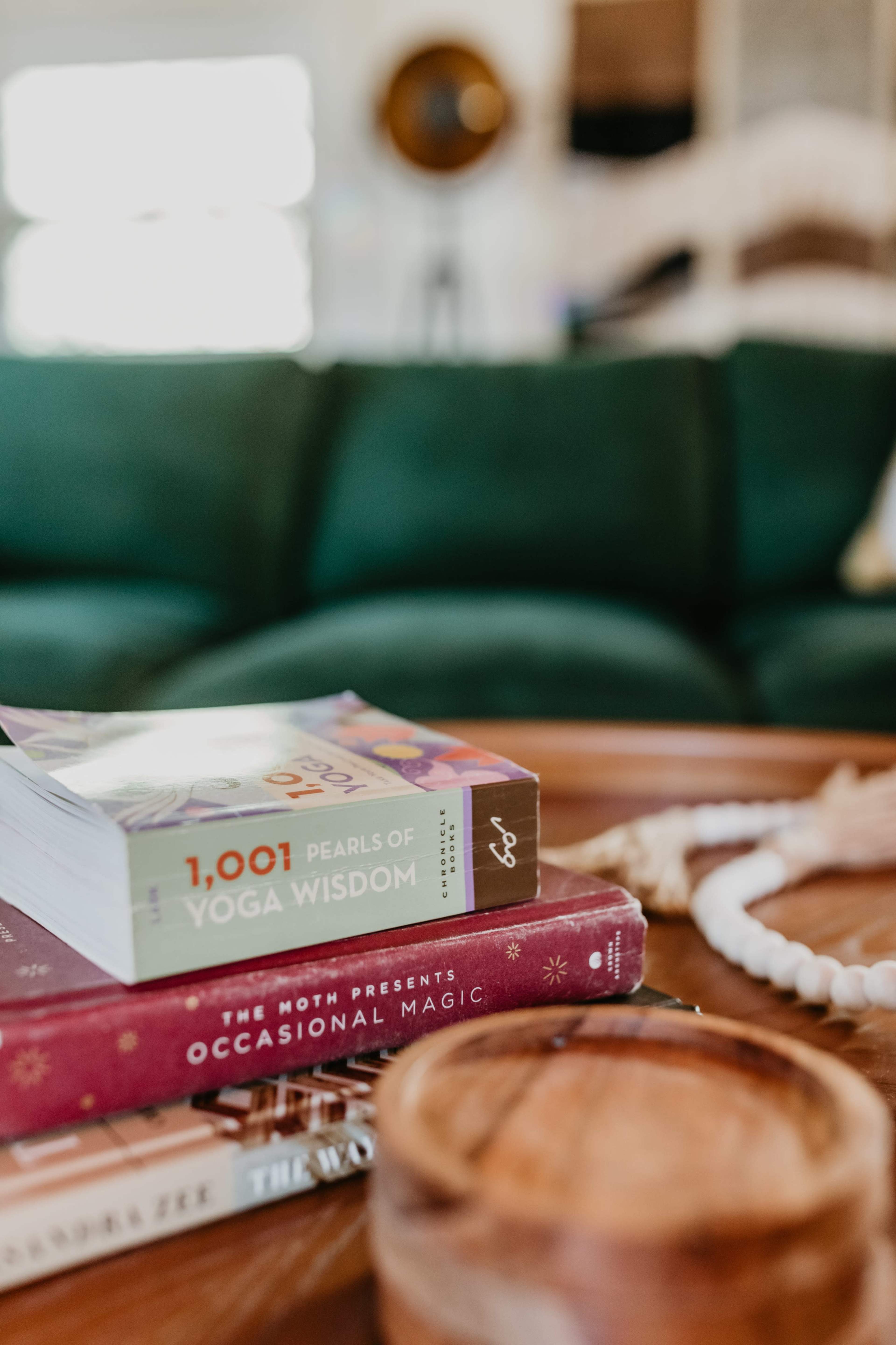 A stack of books, including "1,001 Pearls of Yoga Wisdom" and "Occasional Magic," is displayed on a wooden table in a living room with a green couch in the background.