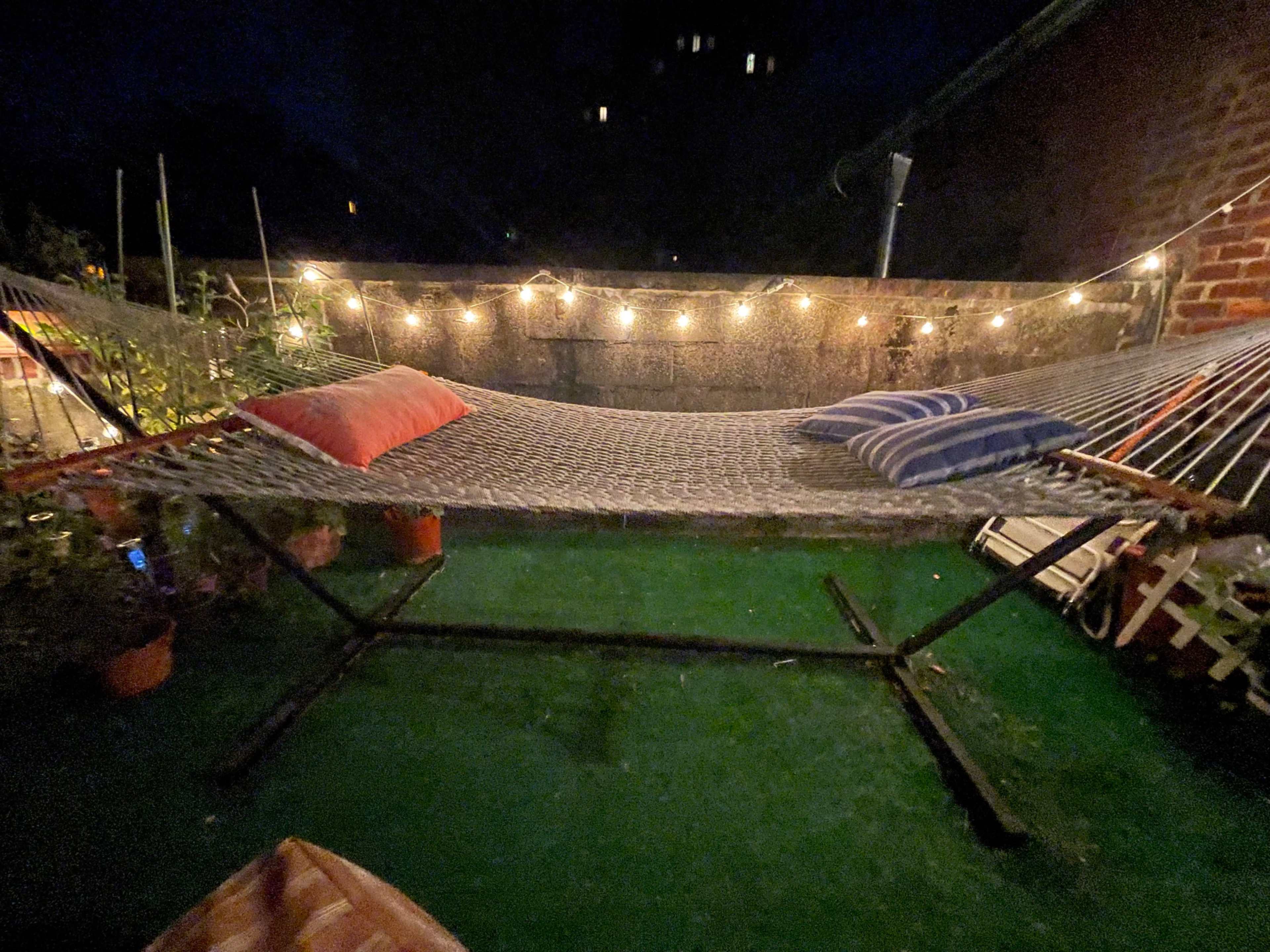 A hammock is set up in a dimly lit outdoor space surrounded by potted plants and string lights.
