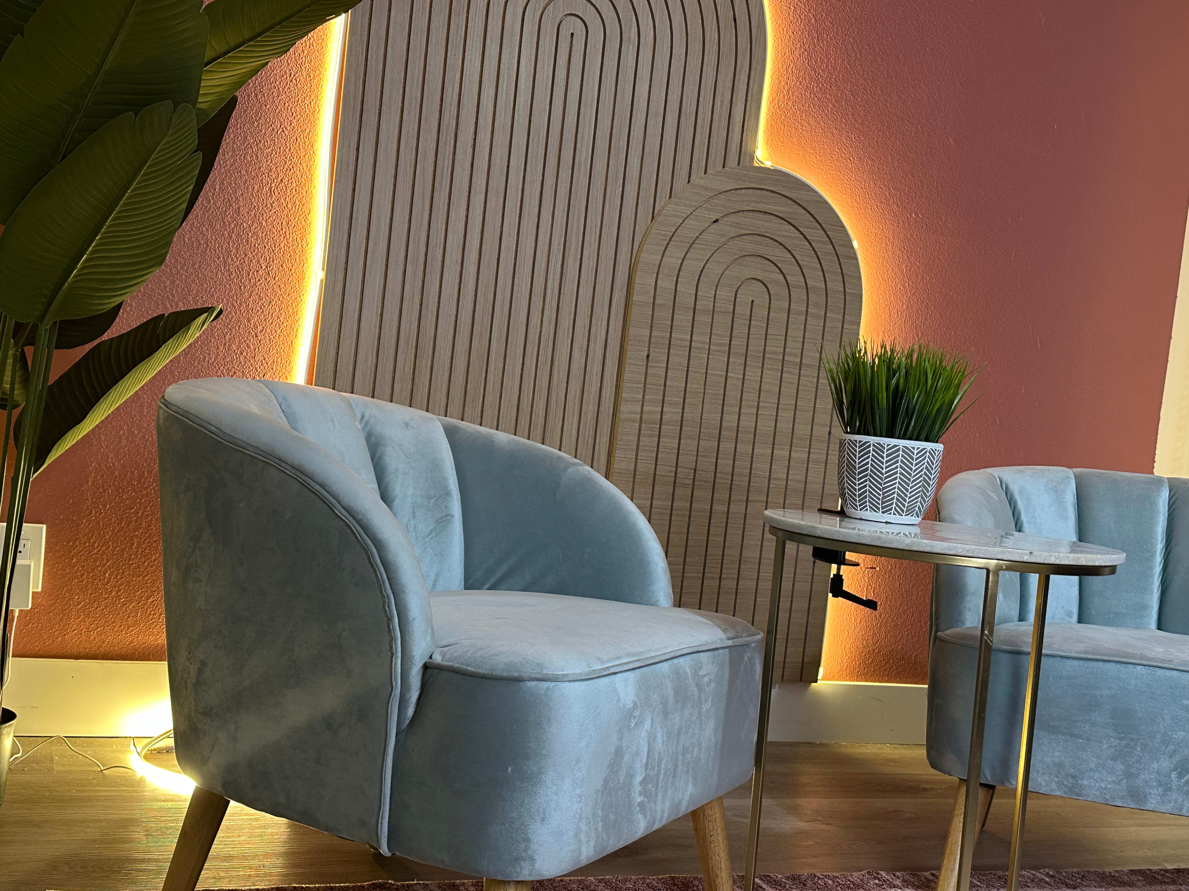 The image shows two light blue velvet chairs next to a small round table with a patterned planter holding a green plant, set against a textured wood panel and softly lit wall.