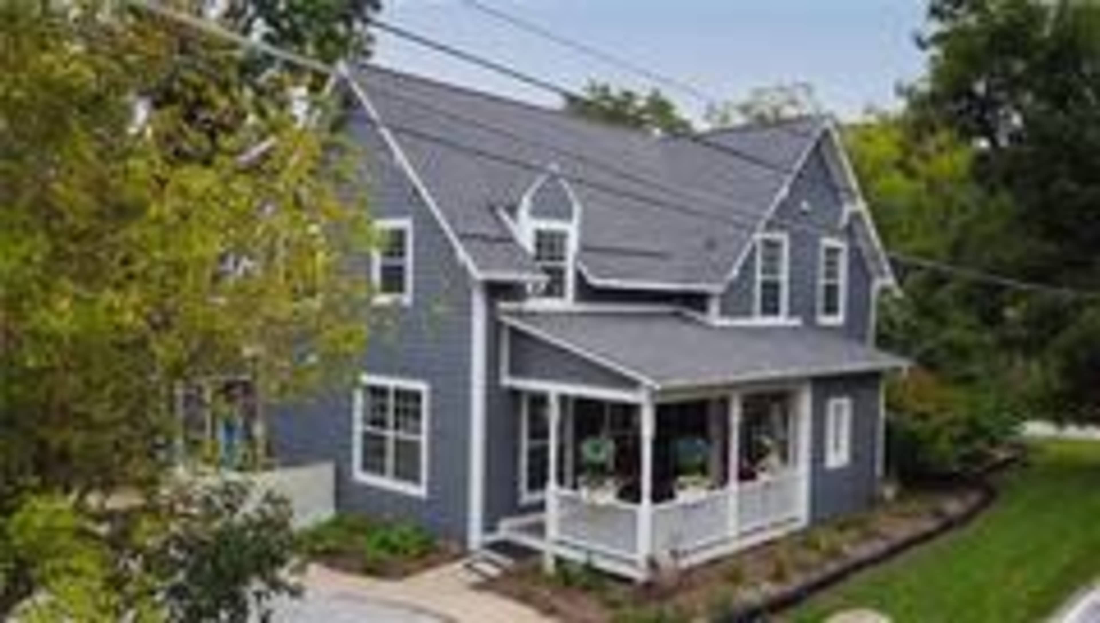 A two-story blue house with a front porch and a landscaped yard is surrounded by trees.