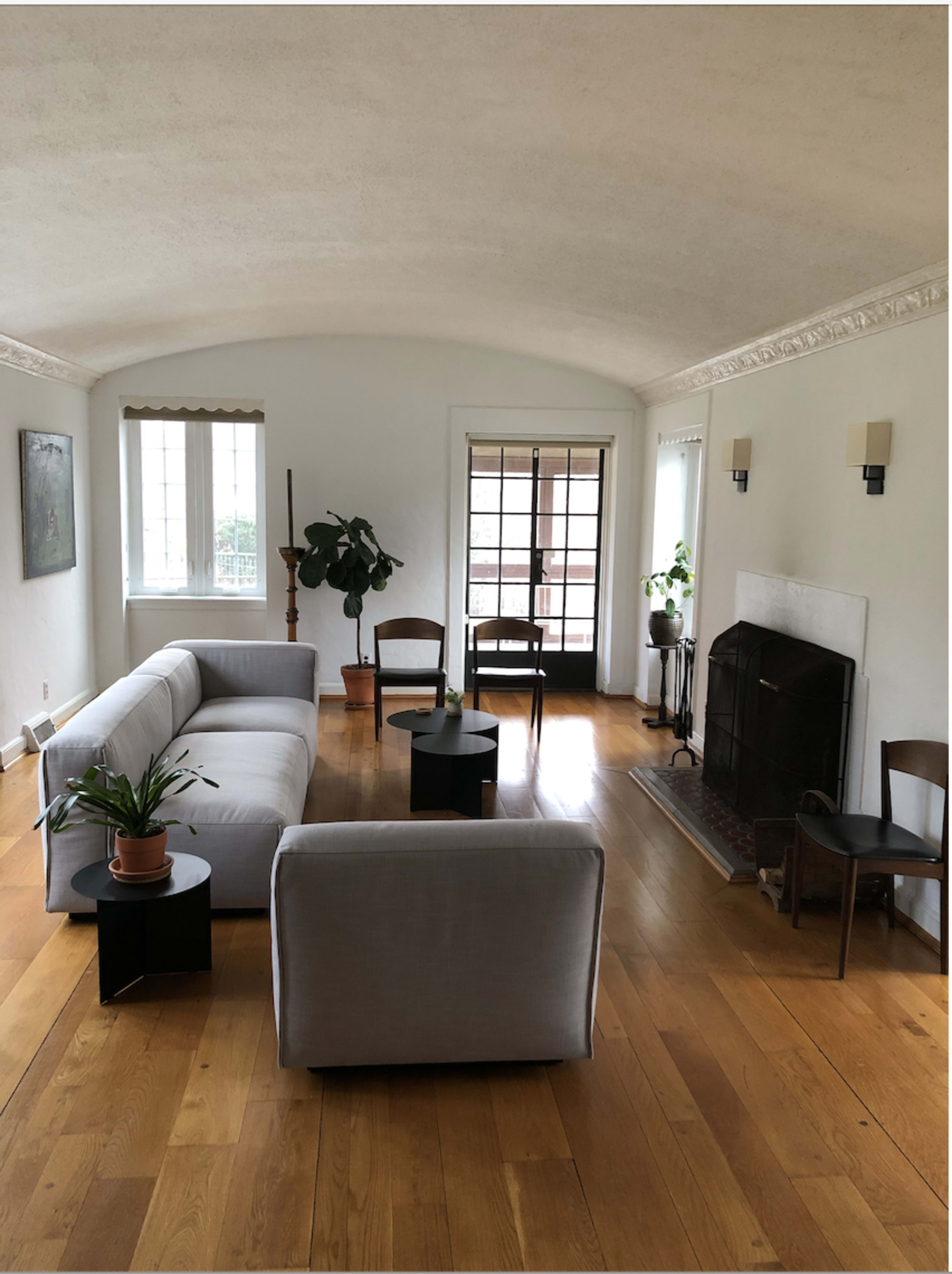 The living room features a light-colored sofa, two black side tables, a fireplace, and several windows, with wooden flooring and minimal furnishings.
