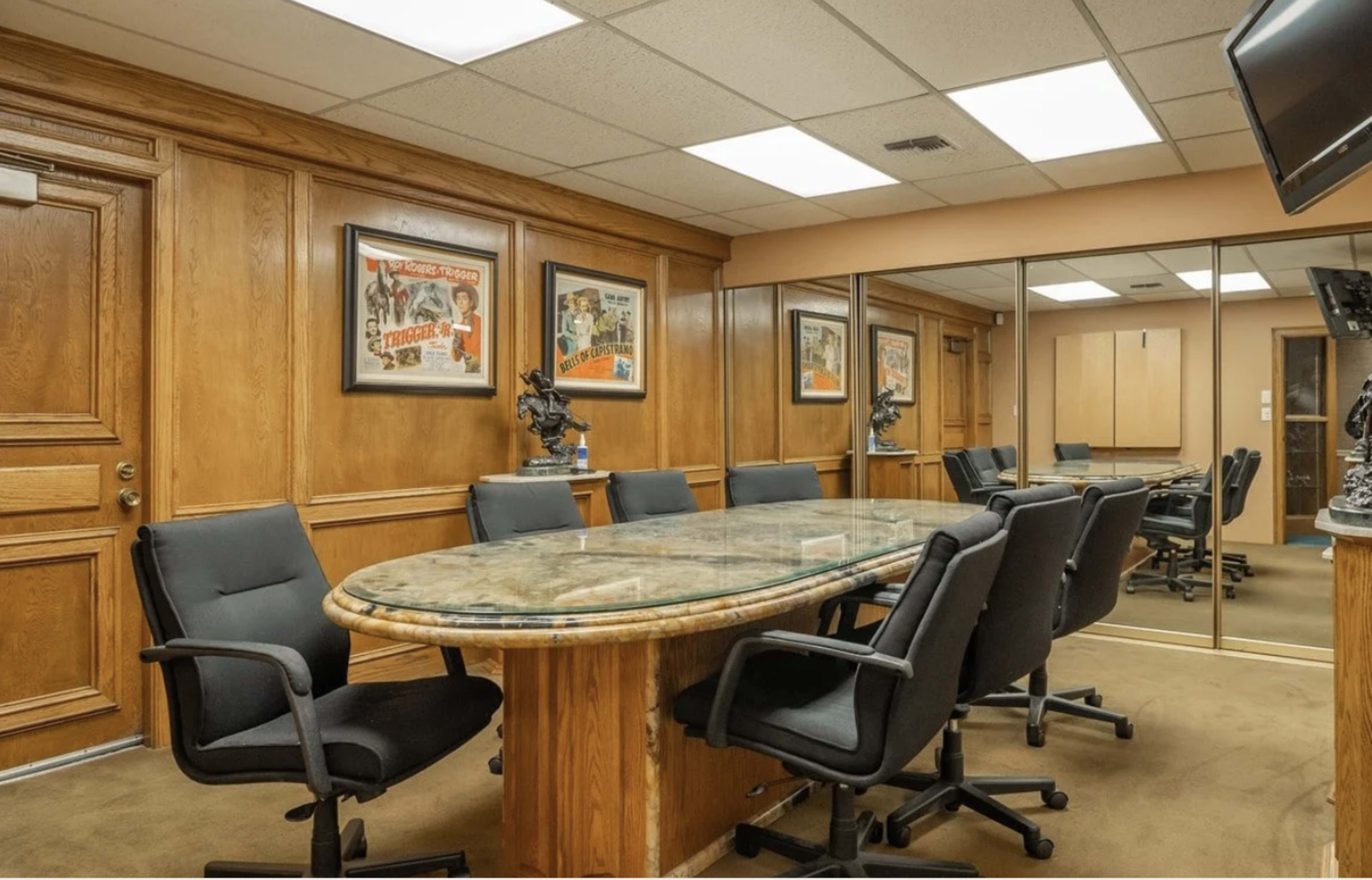 The image shows a conference room with a large glass-topped table surrounded by black ergonomic chairs, wooden paneling on the walls, and framed vintage posters.
